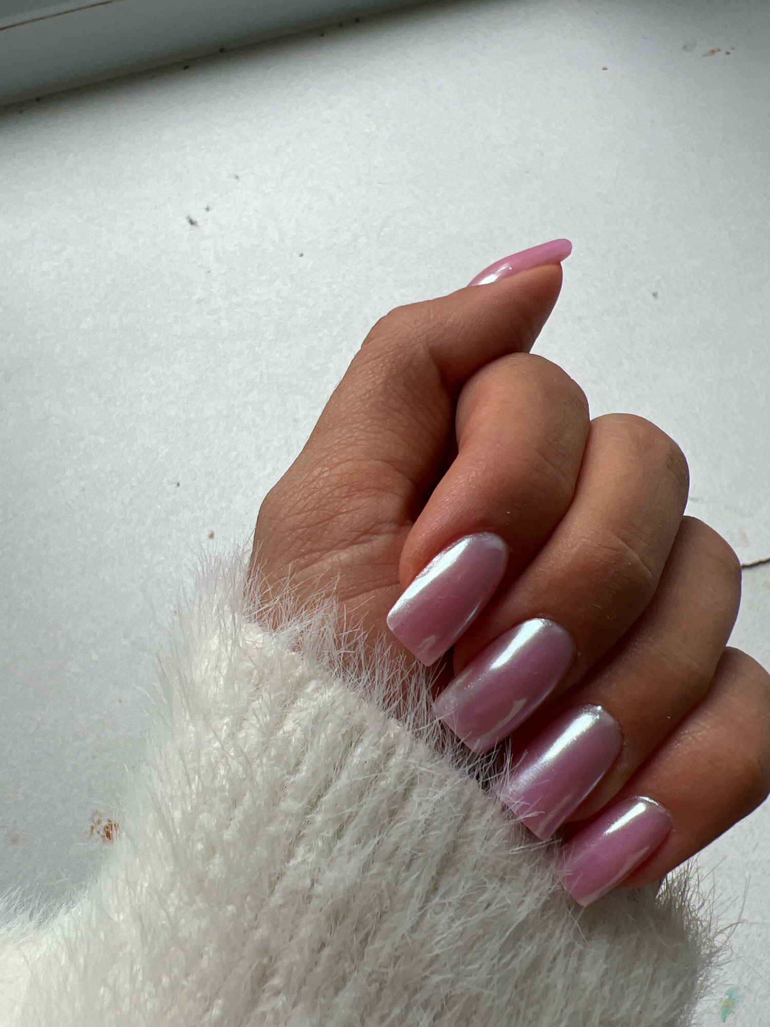 A close-up, high-angle shot captures a left hand adorned with long, square-shaped nails painted a pearlescent pink. The hand is partially covered by a fluffy white sweater. The background is a plain white surface with a few small dark specks and a faint horizontal line across the top. The lighting creates a soft glow on the nails, highlighting their iridescent quality.