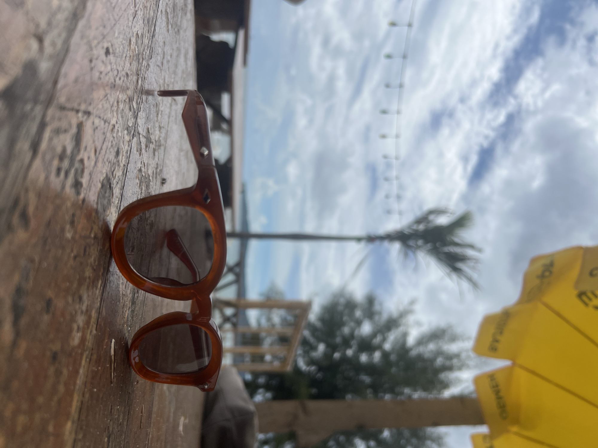 A pair of brown tortoise-shell sunglasses with dark lenses rests on a weathered wooden table. The sunglasses are positioned in the foreground, slightly to the right of center, with the left temple angled away from the viewer. The background is a blurred outdoor scene, suggesting a tropical location, possibly near the coast. A palm tree is visible to the left, and a yellow umbrella with indistinct text is present on the far right. The sky is cloudy with patches of blue. String lights are strung across the upper left of the frame, and a body of water and distant landmass are visible in the mid-ground. The overall impression is a relaxed, beachside setting in Choeng Thale, Thailand.