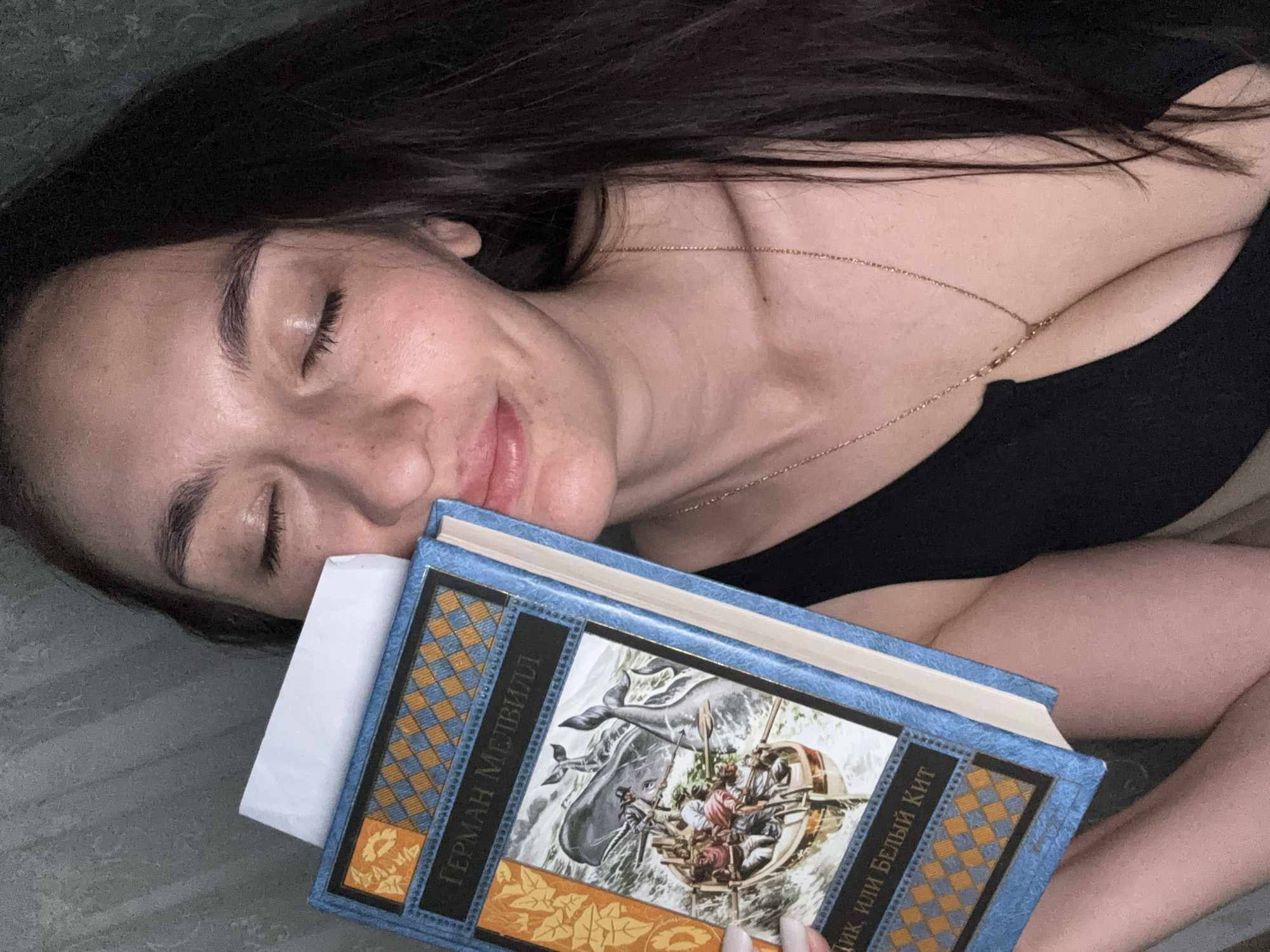 A woman is lying down indoors. Her eyes are closed and she is smiling softly. She has long dark hair and is wearing a black top and a gold necklace. She is resting her head on a book written in Russian. The cover of the book has an illustration of a whaling scene. The woman is fair-skinned, and she has some freckles on her face. The background appears to be a bed or a couch. The location is not specified, but according to the instructions, it is in Sochi, Russia. There is no evidence to support or refute this location.