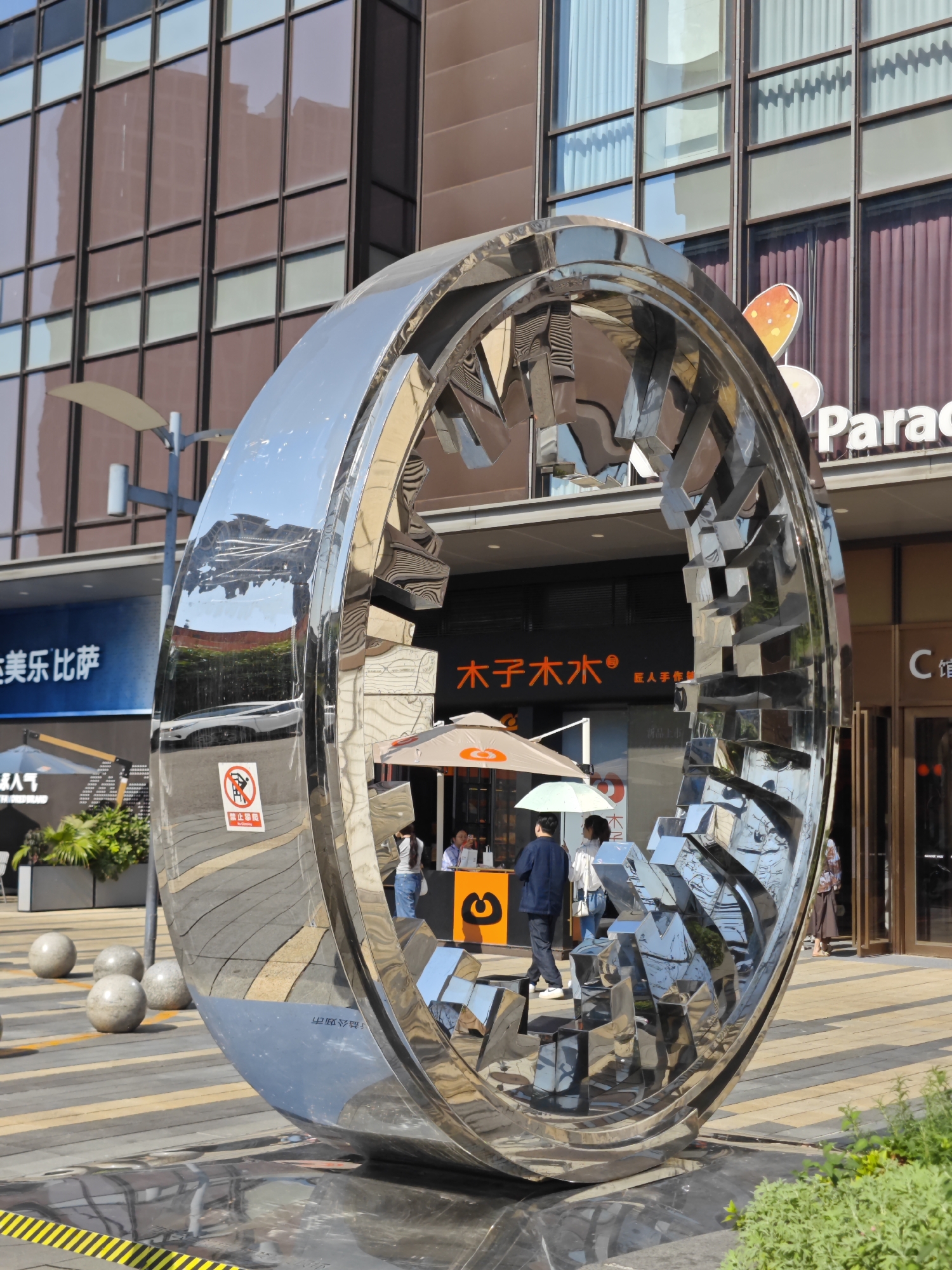 Here is a description of the image:
The image shows a large, shiny, circular sculpture in a public space. The sculpture appears to be made of chrome or stainless steel, and its surface reflects the surrounding buildings and people in a distorted and fragmented manner. The sculpture is positioned on a paved walkway with yellow and black striped markings and round stone or concrete spheres nearby.
In the background, there are modern commercial buildings with storefronts. The signage on the buildings is in both Chinese characters and English, and one sign says "Parad". People can be seen walking near the shops and interacting with the businesses. Some are holding umbrellas.
Based on the presence of Chinese language signage and architectural style, the location is likely a city in China. According to the metadata, this is in Yuzhong District, China. The overall impression is that the sculpture is a piece of public art in a modern, urban setting.