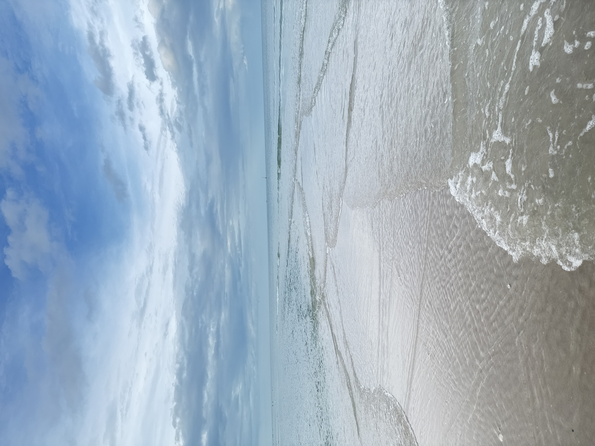 A wide, low-angle shot captures a serene beach scene in Hua Hin, Thailand, under a partly cloudy sky. The foreground is dominated by wet sand, showing intricate ripple patterns left by receding waves, with small shells scattered across the surface. Gentle waves are breaking on the shore, creating foamy white edges that spread across the sand. The midground is occupied by the calm, shallow sea, with distinct lines of incoming waves and lighter patches indicating submerged sandbars. In the distance, the ocean stretches to the horizon, meeting a sky filled with a mix of light and dark gray clouds, interspersed with patches of blue. A lone sailboat is visible on the far horizon, adding a small detail to the vast expanse of the ocean. There are no people or specific identifiable objects in the scene, emphasizing the natural beauty and tranquility of the beach.