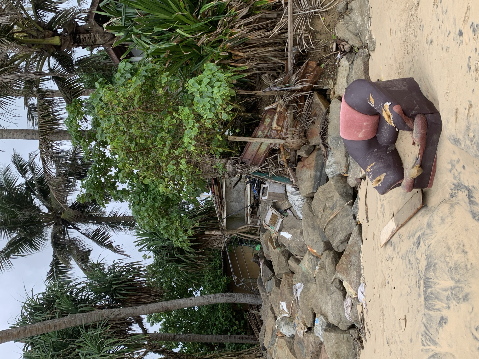 The image depicts a polluted beach scene in Ahangama, Sri Lanka. A dilapidated, dark brown armchair with a pink cushion rests on the sand near a rock barrier. Behind the barrier, there is debris, including what appears to be remnants of a building, amidst dense vegetation, including palm trees. Trash litters the area among the rocks. The sky is overcast.