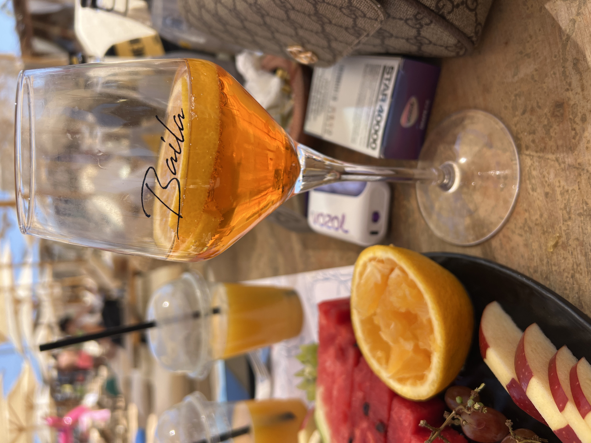 The image shows a close-up of a glass of orange-colored beverage with a slice of lemon and the word "Baila" inscribed on it, placed on a wooden surface. In the background, the scene appears to be a beach resort in Sharm El Sheikh, Egypt, with beach chairs and umbrellas visible. There are also plastic cups with orange juice and a platter of fruits, including watermelon, orange, grapes, and sliced apples. A designer handbag and some electronic devices are also present. The overall impression is a relaxing day at the beach with drinks and snacks.