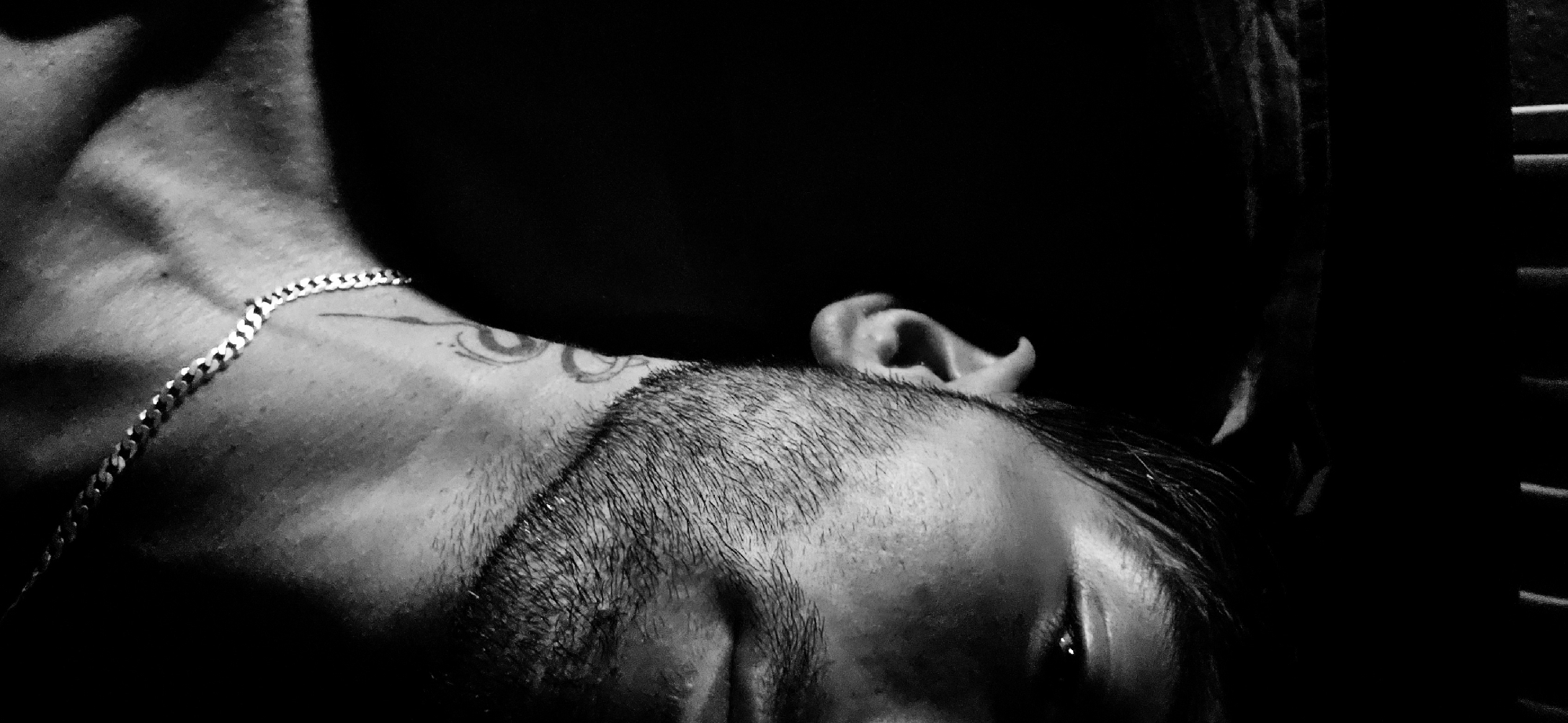 A close-up, black and white, low-angle, right-facing shot features the right side of a person's face and upper chest against a dark background. The person has short, dark hair, stubble on their cheeks and jawline, and a tattoo on their neck. A silver chain necklace is visible around their neck. The lighting emphasizes the contours of the face and neck, with harsh shadows obscuring much of the features. The background appears to be a dark, indistinct space, possibly indoors.