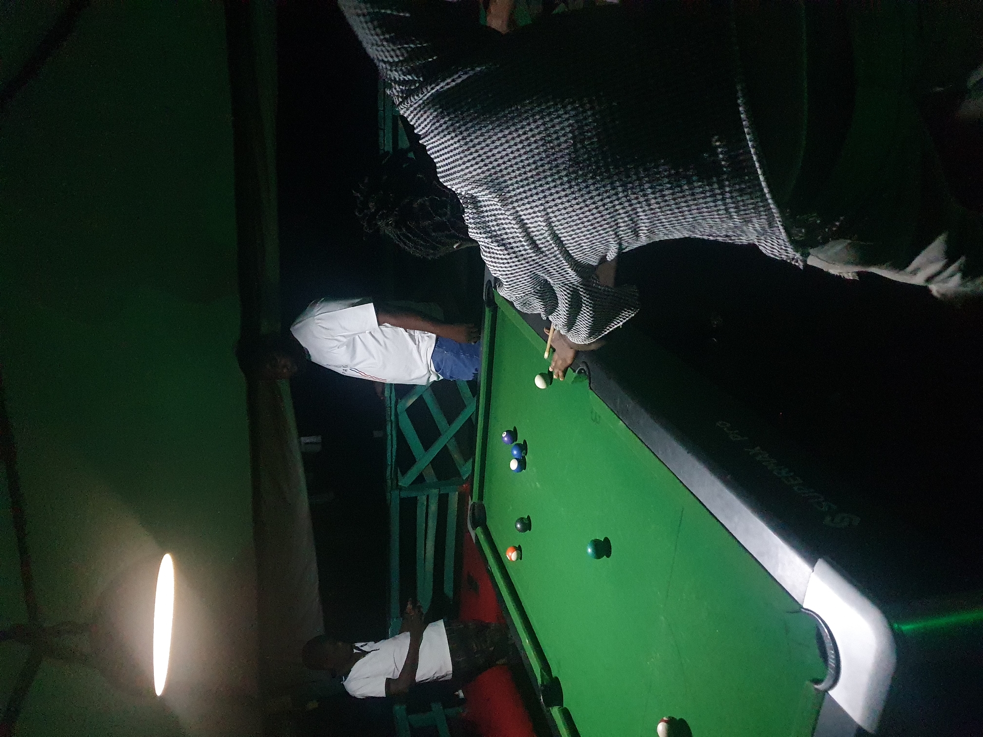 A dark, outdoor, possibly covered, recreational area in Lokoja, Nigeria, features a pool table under the illumination of a bright, oval overhead light. Three men are present. One man, on the far left, stands near the table, wearing a white polo shirt and camouflage shorts, holding a cue stick. Another man, in the center background, stands behind the table, wearing a white t-shirt and blue jeans. The third man, in the foreground and facing away from the camera, is actively playing pool, dressed in a patterned long-sleeved shirt and light-colored pants. Several pool balls are scattered across the green felt surface of the table. A green railing is visible behind the players. The overall atmosphere is casual and dimly lit, suggesting an evening activity.