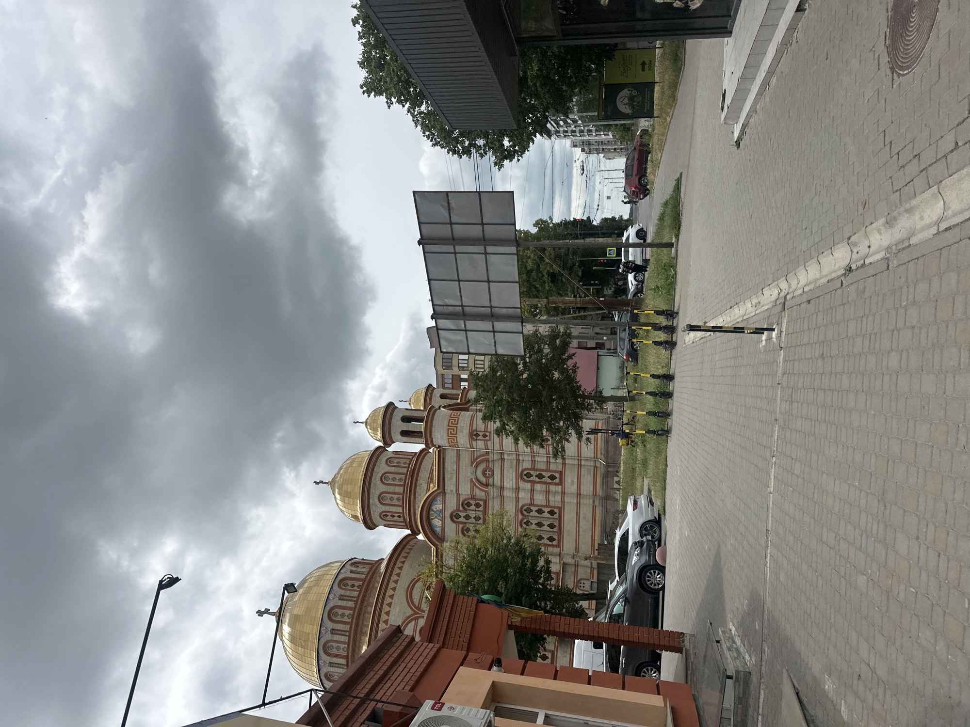 The image shows a street scene in Ternopil, Ukraine, with a prominent Orthodox church featuring golden domes in the background. To the left, a brick building with an air conditioning unit is visible. Several cars are parked along the street, and a row of electric scooters stands near the curb. The street is paved with cobblestones, and a sidewalk runs alongside it. The sky is overcast with dark, heavy clouds, suggesting impending rain. In the distance, a billboard, trees, and apartment buildings can be seen.