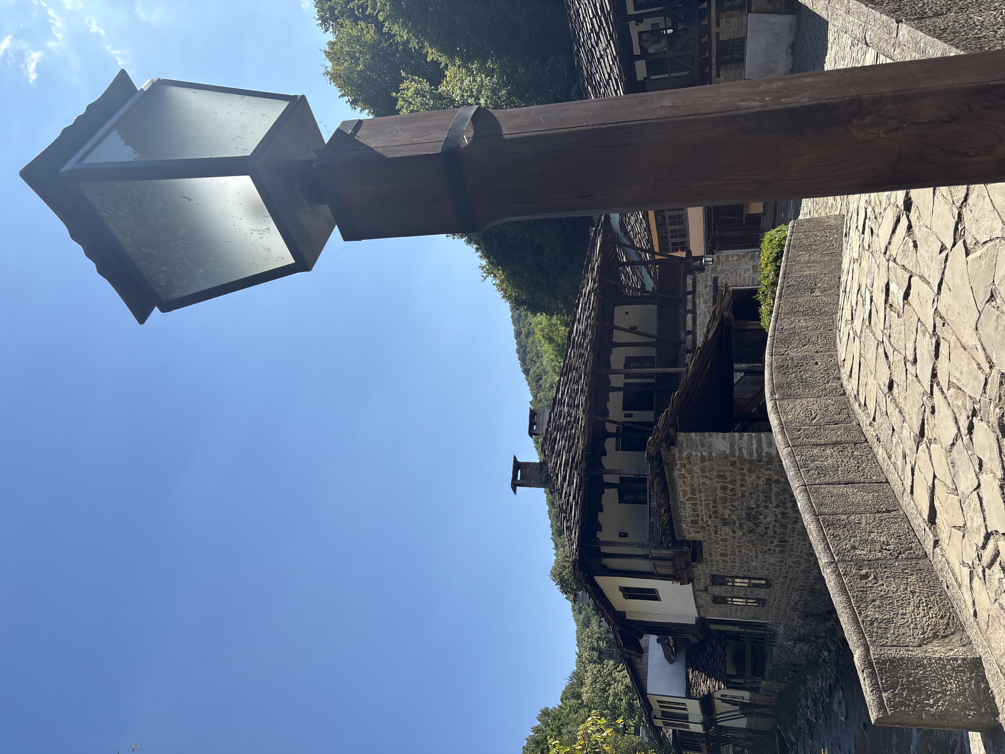 A vintage lantern with a weathered glass shade stands on a wooden post in the foreground, casting a diffused light. Behind it, a rustic stone bridge arches over a body of water, its surface paved with irregular stones. On the far side of the bridge, traditional stone and timber houses with dark tiled roofs line a cobblestone street. The architecture suggests a historical or rural setting, with overhanging balconies and stone chimneys. Dense green foliage and trees form a backdrop on a sloping hillside under a clear blue sky. The scene is bathed in bright daylight, with shadows indicating a sunny day. The location is identified as Unknown, Unknown.