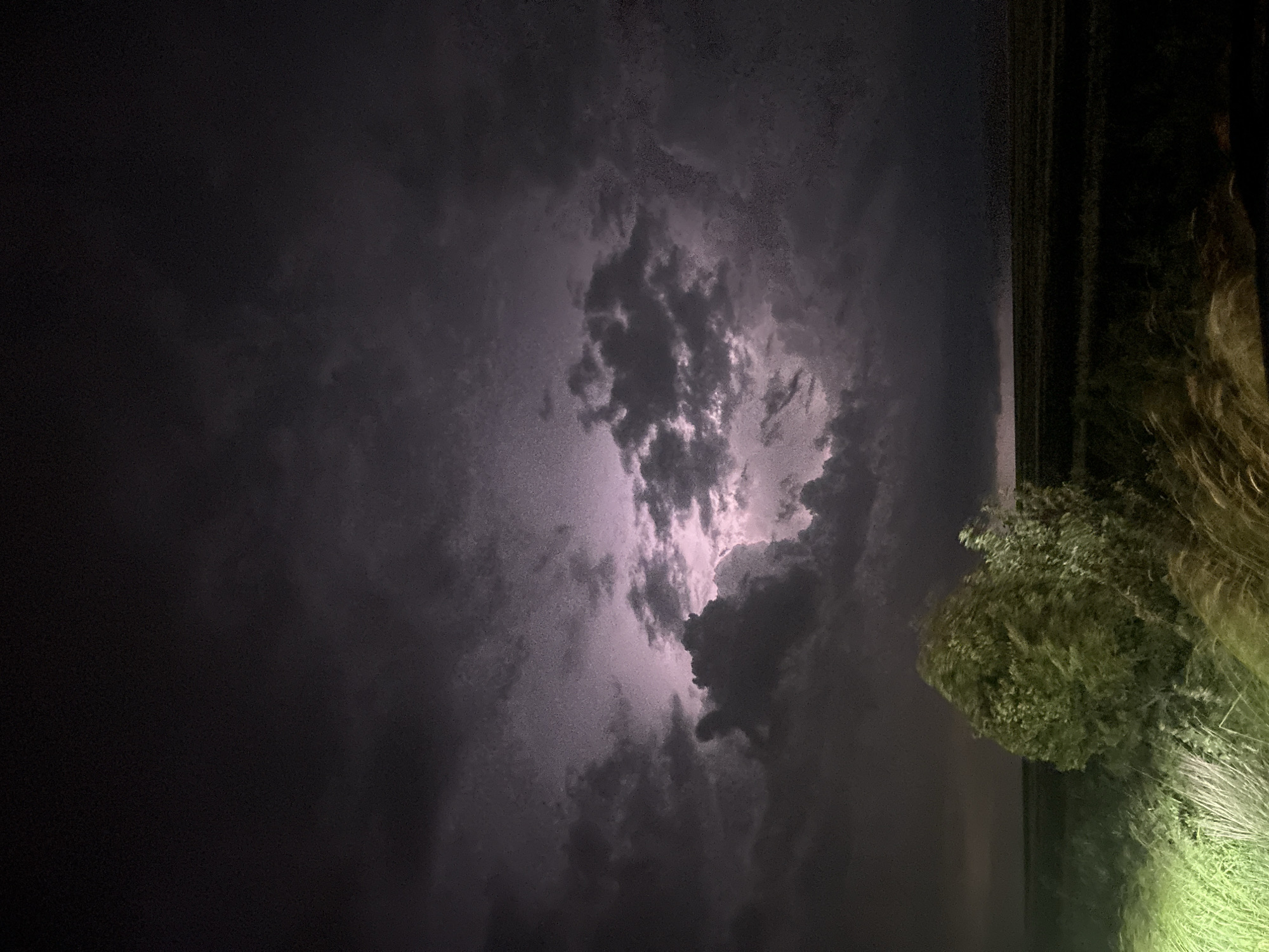 The image depicts a nighttime scene of a stormy sky. Heavy, dark clouds dominate the view, with a brighter area suggesting lightning or other electrical activity within the cloud cover. The right side of the image includes some foliage, potentially bushes or trees, illuminated by an unseen light source. A dark band of land or a distant structure is visible near the top of the frame. There is no evidence to verify the location in the city of Burgas, Bulgaria. The primary subjects are atmospheric conditions and vegetation at night.