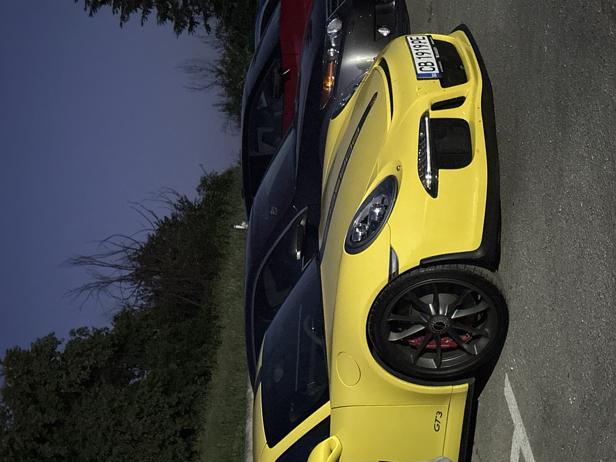A bright yellow Porsche GT3 is parked on asphalt in front of a line of other cars, including a dark colored sedan and a red vehicle, under a dark blue sky with trees in the background. The yellow Porsche is in the foreground, facing right, with its front wheel visible, along with its license plate which reads "CB 1919PE". The car has a black stripe running down the hood. The environment suggests an outdoor parking area in Arbanassi, Bulgaria, likely during dusk or early evening. No people are visible in the frame.