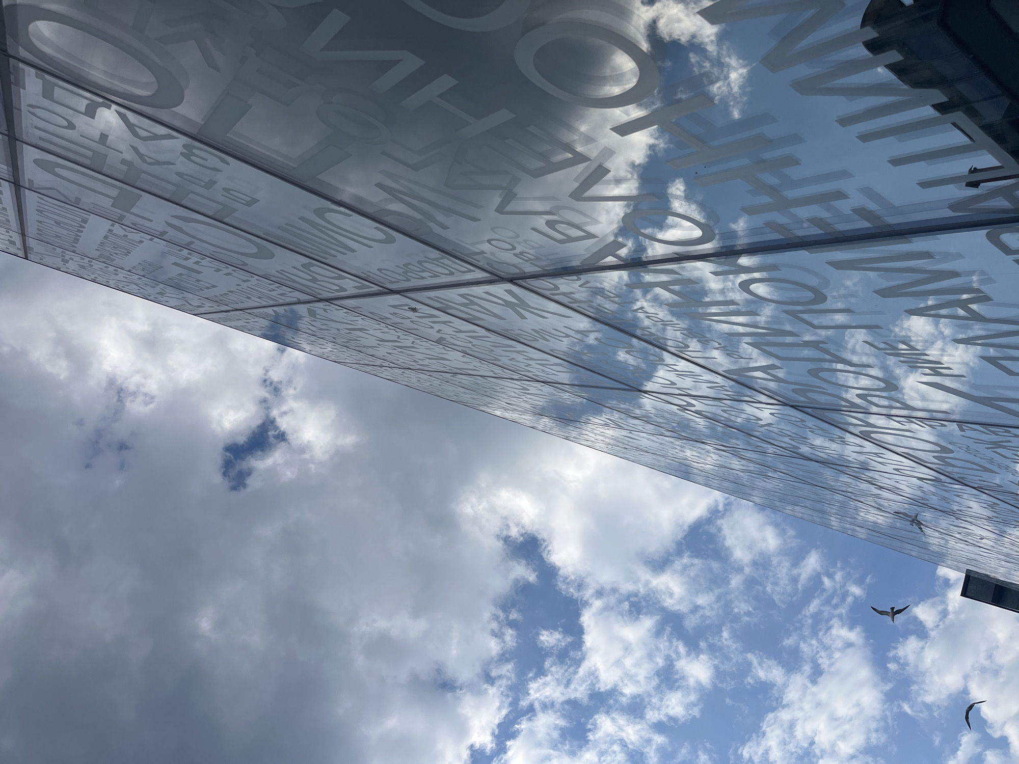 A low-angle shot shows a building facade with white lettering on a glass surface reflecting the sky. The sky is mostly blue with scattered white and gray clouds. There are three birds flying in the sky, positioned in the lower right of the frame. The building appears to be in an urban location, likely the city of Burgas, Bulgaria.