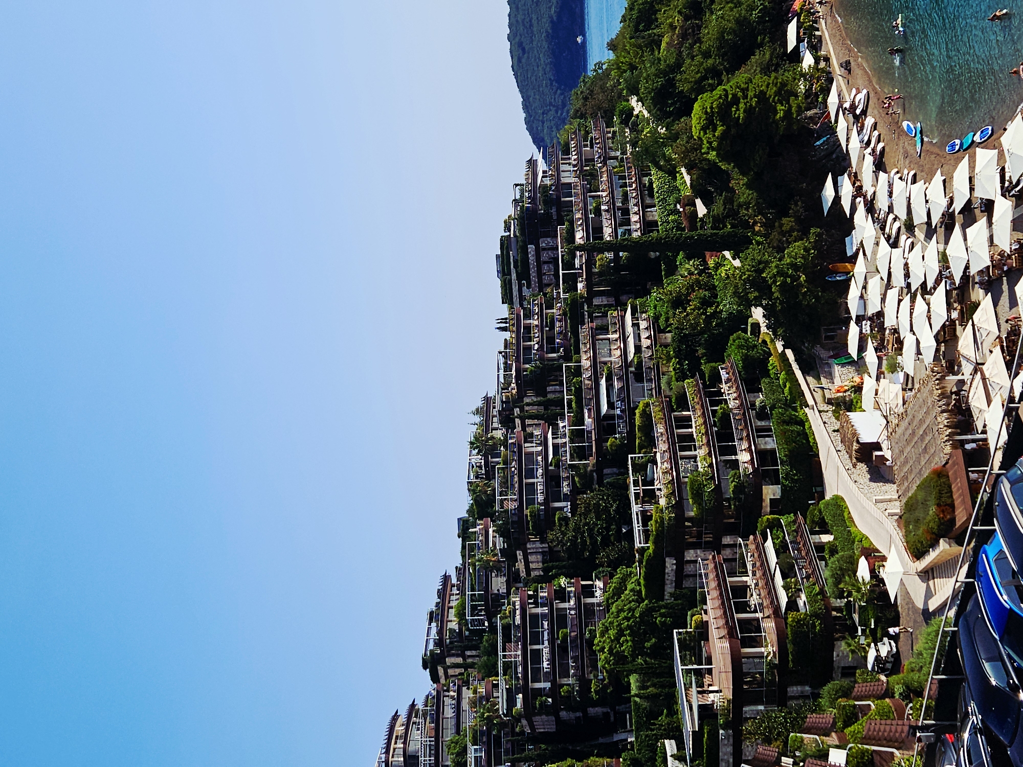 The image depicts a luxurious resort complex built into a hillside overlooking the Adriatic Sea, located in Budva, Montenegro. The architecture features multi-tiered buildings with numerous balconies and terraces, heavily integrated with lush greenery, creating a terraced effect. The resort appears to have extensive amenities, including a beach area with numerous white umbrellas and loungers, several people enjoying the water, and a small fleet of surfboards and paddleboards. A road winds through the resort, with a few golf carts and parked cars visible. The sky is a clear, bright blue. In the distance, a verdant, tree-covered hill slopes down to the sea, with a lone boat visible on the water.