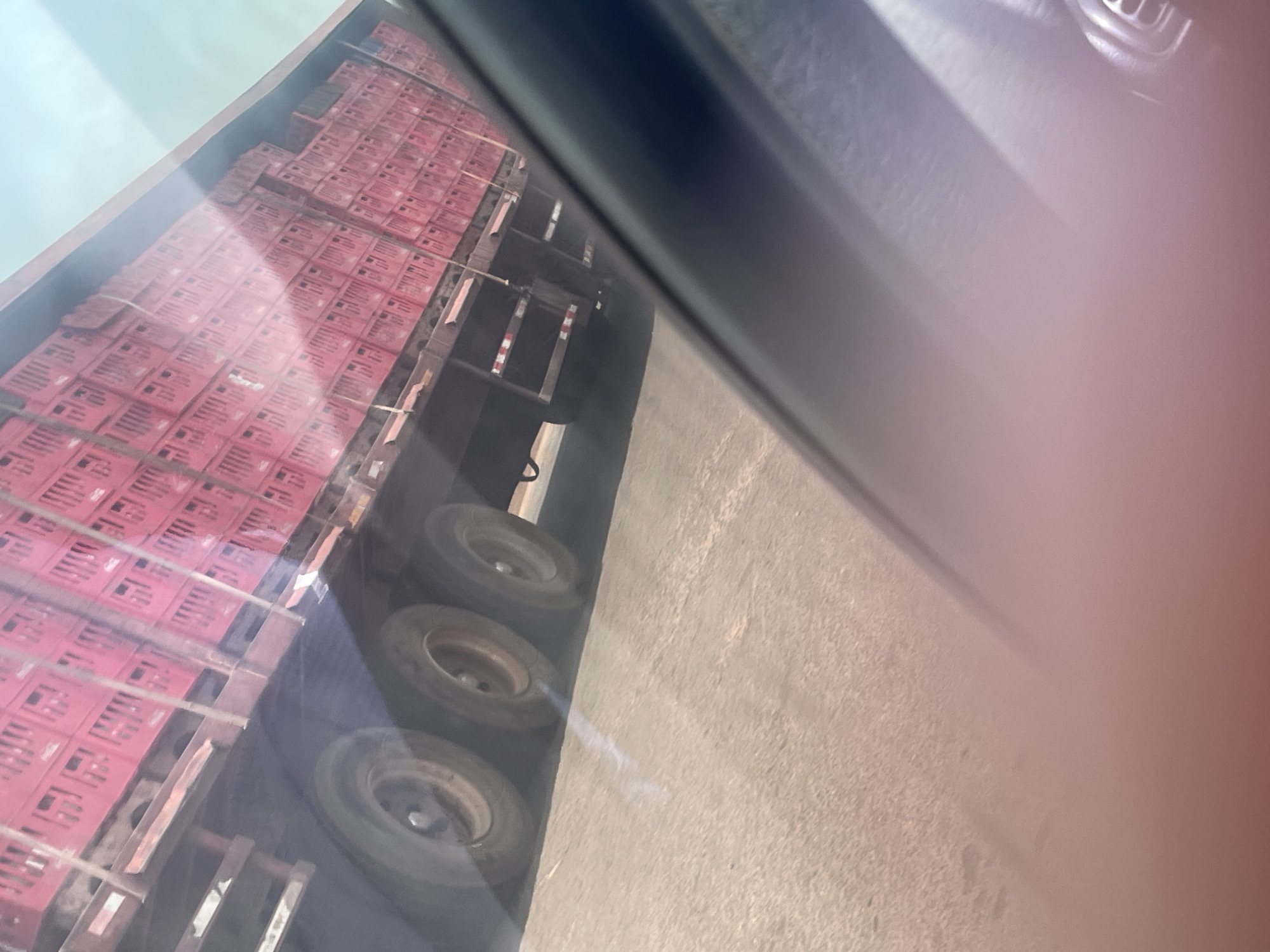 The image, captured in Keffi, Nigeria, presents a view from inside a vehicle, looking out through a side window. The right and top portions of the frame show blurred, dark shapes, indicative of the car's interior, possibly a door frame or dashboard. The central subject outside is a large truck or trailer heavily loaded with numerous red plastic crates.
These crates are stacked high, forming multiple rows and columns along the length of the truck bed, suggesting a significant quantity of goods being transported. While the specific contents of the crates are not visible, their uniform appearance and large volume imply commercial logistics, possibly for agricultural produce or other commodities. Several robust wheels of the truck are visible in the lower-left area, consistent with a heavy-duty transport vehicle.
The truck is positioned on what appears to be a road or a roadside, with a light-colored ground surface, likely asphalt or concrete, extending beneath it. The upper-left corner reveals a bright sky, indicating that the photo was taken during daylight hours, although precise weather details are obscured by reflections and haze on the car window. No people are visible in the frame, and the activity depicted is primarily the presence of the loaded truck, suggesting it is either in transit or parked. Indistinct textual markings are present on some of the red crates, but they are too blurry to be deciphered.