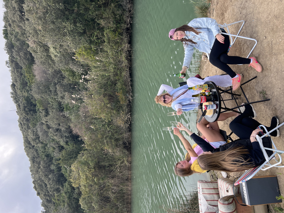 Four females are positioned outdoors beside a calm, greenish-blue body of water, backed by a densely vegetated hillside, under an overcast sky. The location is in the city of Unknown, Unknown. The ground is dry, exposed earth.
From left to right, seated around a small, black, round folding table:
1. A female with long brown hair, wearing a black top and black leggings, sits on a white and blue striped folding chair with her back partially to the camera. A dark grey portable speaker is situated to the left of her chair.
2. A female with light brown hair, wearing a yellow top and pink shorts, is seated, extending a champagne flute forward.
3. A female with blonde hair and sunglasses perched on her head, wearing a white long-sleeved top and a light blue shawl, is seated, extending a champagne flute forward and smiling towards the camera.
4. A female wearing a pink baseball cap, a light blue denim jacket, black leggings, and pink sneakers, is seated on a white folding chair, extending a green beer bottle forward and smiling towards the camera.
The central table holds one Coca-Cola bottle, one Bavari beer bottle, three other unidentified beverage bottles, a small red container, a knife, and a white-wrapped food item. On the far left, a striped upholstered object resembling furniture components lies on the ground. The far bank of the water body is characterized by tall reeds and dense green shrubbery, which transitions into a steep hillside covered with evergreen trees.