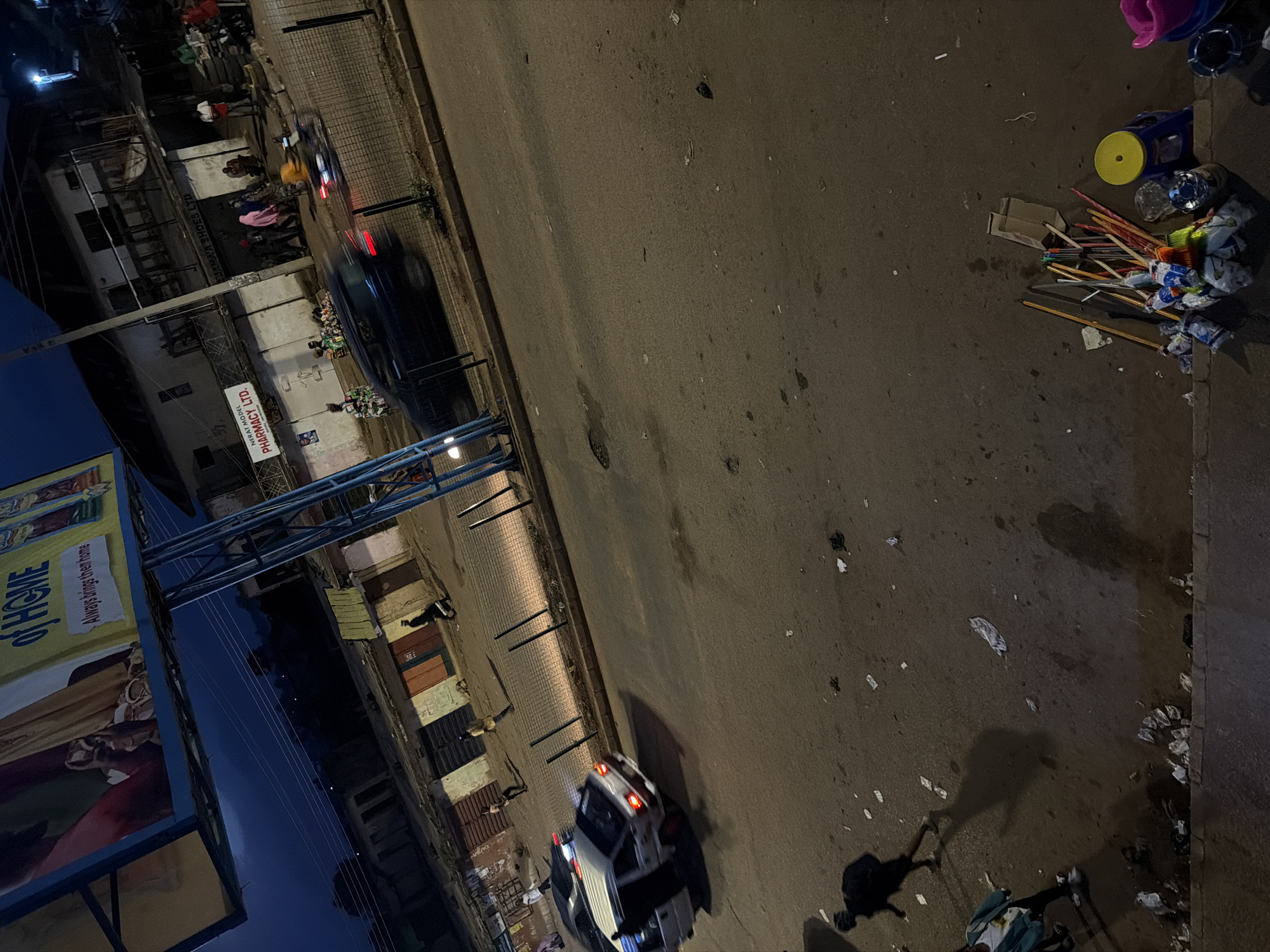 The image depicts a street scene at dusk or night in Bukuru, Nigeria. A billboard advertising "OF HOME" is prominently displayed, with smaller text reading "Always brings them home." Below the billboard, a busy street is visible, with several buildings lining one side. One building has a sign that reads "NERAT MODEL PHARMACY LTD." and another that says "JAMAVA SHOES LTD." People are walking and gathered on the sidewalk, and several vehicles are on the road. One car, a white van, is in motion, with its doors open. Another dark-colored car is also moving, showing motion blur. There are streetlights visible in the distance. The ground is paved and appears somewhat worn, with scattered debris. In the foreground, a collection of brooms and cleaning supplies are neatly arranged near a yellow-topped stool. The overall atmosphere suggests a commercial area that is still active at this late hour.