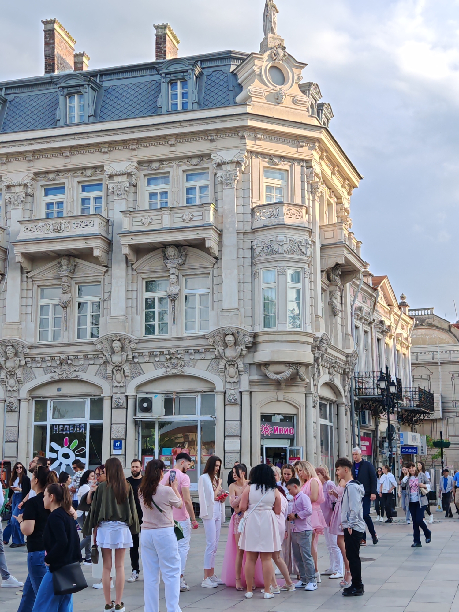 The image depicts a gathering of people on a city street in Ruse, Bulgaria. Dominating the background is a grand, ornate building with elaborate architectural details, including sculptures, balconies, and a mansard roof. The building appears to be a historical structure. The foreground is populated by a group of people, many of whom are dressed in shades of pink or white. Some individuals are interacting and taking photos, suggesting a special occasion or event. Other pedestrians are casually walking by. Storefronts with signage in Cyrillic are visible on the ground floor of the building. Streetlights and other buildings can be seen in the background. The overall atmosphere is that of a bustling urban environment.
