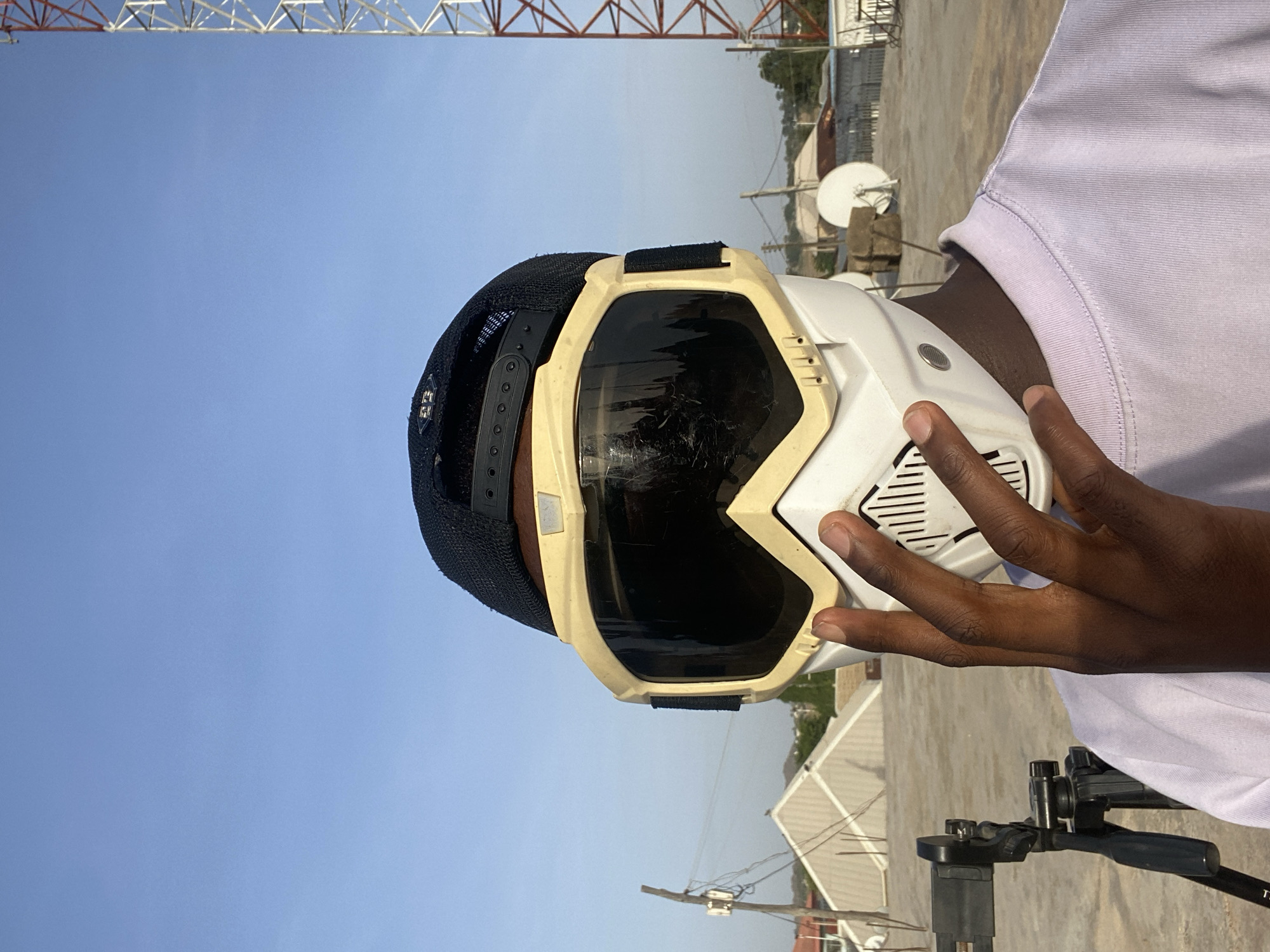 An outdoor, portrait-oriented shot depicts an individual from the chest up, set against a clear blue sky. The person, with dark skin, is wearing a black mesh baseball cap backward, beneath a white, full-face protective mask or goggles. The mask features dark, reflective lenses, covering the eyes, nose, and mouth area, with vented sections on the sides. A light purple short-sleeved top is worn by the individual. A hand is visible, holding or adjusting the side of the mask.
The background is dominated by a clear blue sky, indicating bright, sunny daytime weather. Spanning the top edge of the frame is a large red and white metal lattice tower, likely a communication or electricity pylon. Below the horizon, the scene suggests an elevated concrete surface, possibly a rooftop. Distant structures include buildings with light-colored roofs, a white satellite dish, and a wooden utility pole with wires. In the bottom right corner, a black camera tripod with a phone mount is partially visible. No discernible text is present on the clothing or gear. The overall image captures an individual in protective gear outdoors in Bukuru, Nigeria, potentially preparing for an activity or filming.