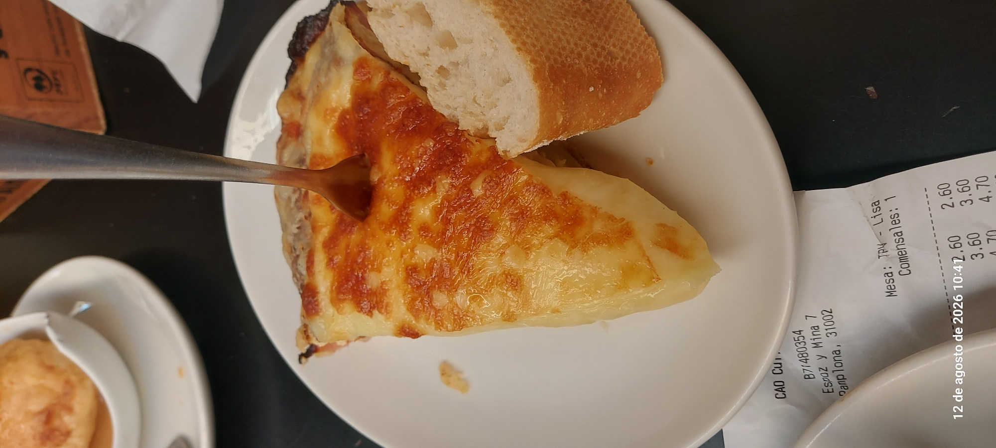 A close-up, slightly overhead shot captures a slice of Spanish tortilla (potato omelet) on a white plate, with a spoon inserted into it. A piece of bread is positioned to the right of the tortilla. The background is dark, likely a tabletop. Partially visible at the bottom of the image is a receipt from "CAO Cuti" in Pamplona, Spain, located at "Espoz y Mina 7, Pamplona, 31002." The receipt also indicates "Mesa: TPV - Lisa," "Comensales: 1," and a date and time stamp of "12 de agosto de 2026 10:41," followed by various prices. The scene suggests a casual dining setting in Pamplona.