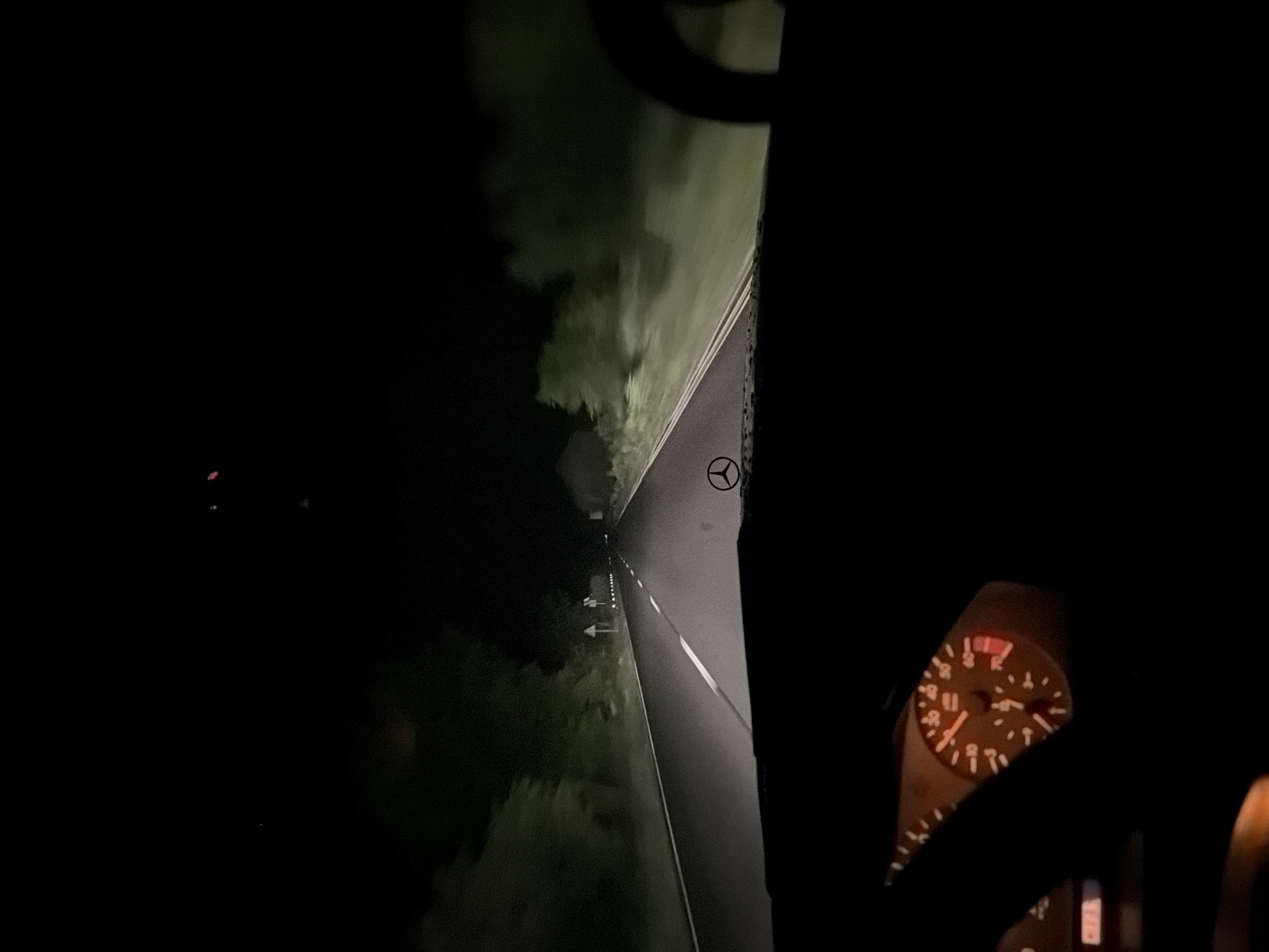 The image depicts a night-time view from inside a vehicle traveling on a road. The road appears to be paved and has lane markings indicating the direction of travel. The surrounding environment is dark with vegetation visible on the sides of the road. An instrument panel with illuminated gauges is visible in the lower right portion of the frame. A steering wheel is visible. Based on the direction signs, it is likely near Blagovo, Bulgaria, but the specific location is not discernible from the image. There is no identifiable activity or interaction taking place.