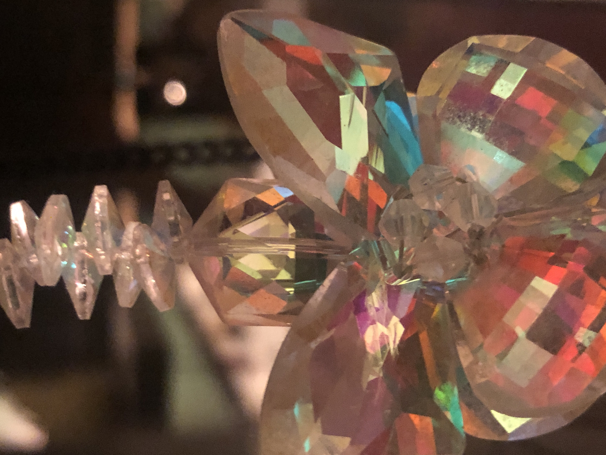 A close-up, shallow depth of field photograph captures a faceted crystal ornament hanging against a dark, blurred background. The ornament is shaped like a flower, with layered petals that refract light, creating a spectrum of colors including pink, orange, yellow, green, blue, and purple. A series of smaller, flat, stepped crystal beads are strung above the flower-shaped ornament, also catching and reflecting light. The background is indistinct, appearing as dark brown and green tones, with a small, out-of-focus circular light source visible on the right side. The overall impression is one of sparkle and luminescence.