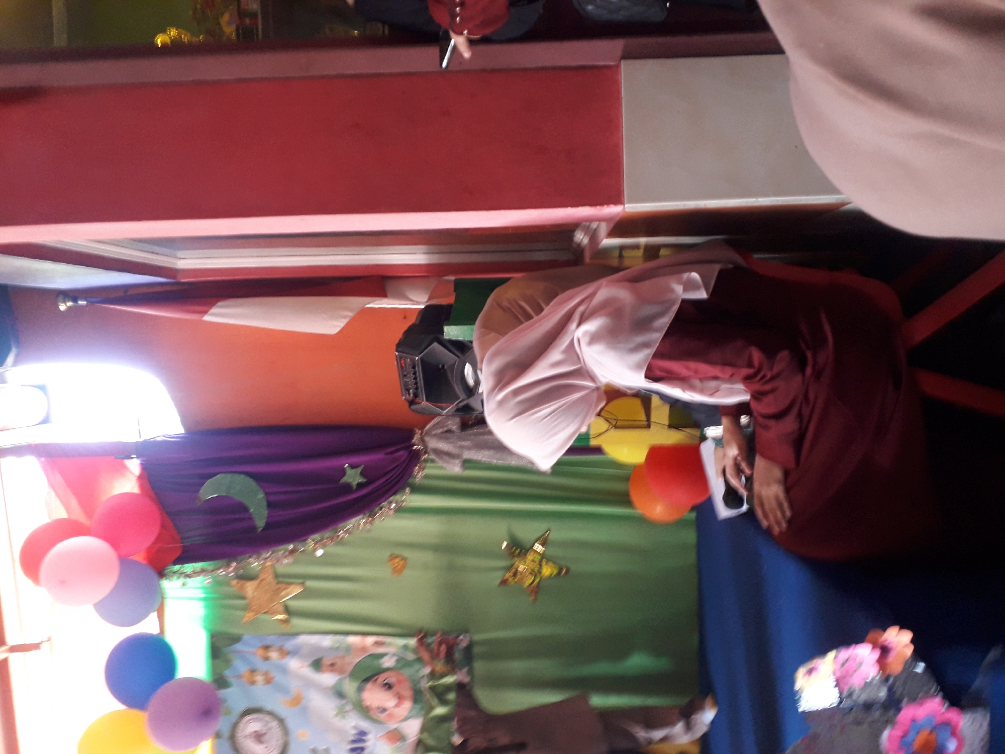 In the city of Cankuang, Indonesia, a dimly lit room is decorated for a festive occasion, possibly an Islamic celebration given the crescent moon motif. The walls are painted in vibrant hues of orange and red. A prominent red pillar divides the space. In the foreground, a person wearing a maroon dress and a light pink hijab sits on a red chair, their hands resting on a table. They appear to be holding a microphone and a sheet of paper. To the left, a green curtain serves as a backdrop, adorned with a large green crescent moon and a gold star. A banner featuring a cartoon character is also visible. Above the curtain, a cluster of colorful balloons – yellow, blue, lavender, and pink – floats, with more balloons, red and yellow, visible lower down. A red and white Indonesian flag hangs to the right of the frame. In the background, part of a shelf with trophies or awards is visible. The overall impression is of a lively gathering or event.