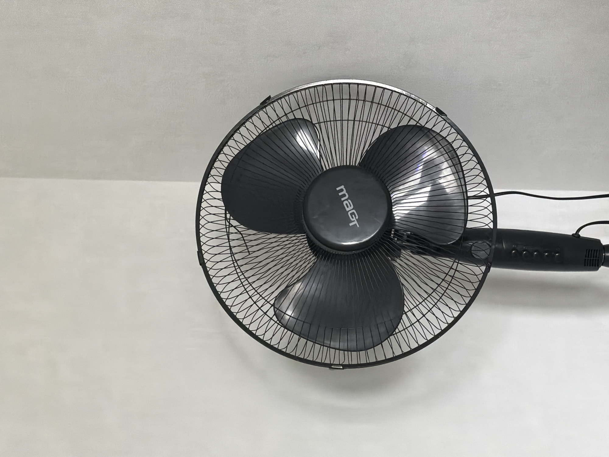 A black floor fan stands in a corner where two light gray walls meet. The fan has a circular wire grill and three dark gray blades. The brand name "magr" is visible on the central hub of the fan, printed in white text that appears to be mirrored. The fan's stem and base are also black, with visible buttons on the stem. A black power cord extends from the base. The environment appears to be indoors, likely a room, with plain walls and no other objects in the immediate vicinity. The lighting suggests an ambient indoor light source. This scene is unlikely to be identifiable to a specific location in Novosibirsk, Russia without further context or identifying landmarks.
