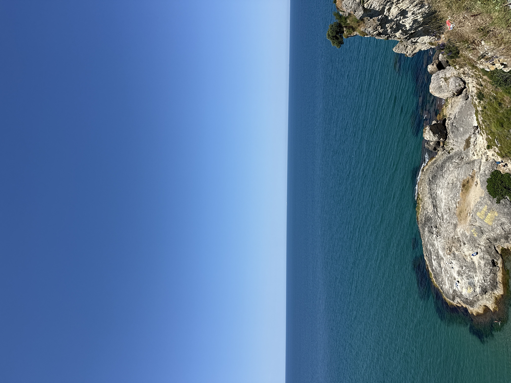 An aerial view captures a bright, clear day with a vast expanse of turquoise water meeting a cloudless, deep blue sky. In the foreground, rugged grey rocks jut out into the sea, with a few small figures scattered across them, some appearing to be sunbathing or relaxing. The coastline is rocky and sparsely vegetated with dry grasses and a few scattered green bushes. To the right, a dramatic cliff face rises from the water, adorned with patches of green vegetation and a single tree clinging to its edge. The image appears to be taken from a high vantage point overlooking a coastal area, likely the Black Sea coast near Şile, Turkey, given the distinct rock formations and vegetation characteristic of the region. The water is calm with gentle ripples.
