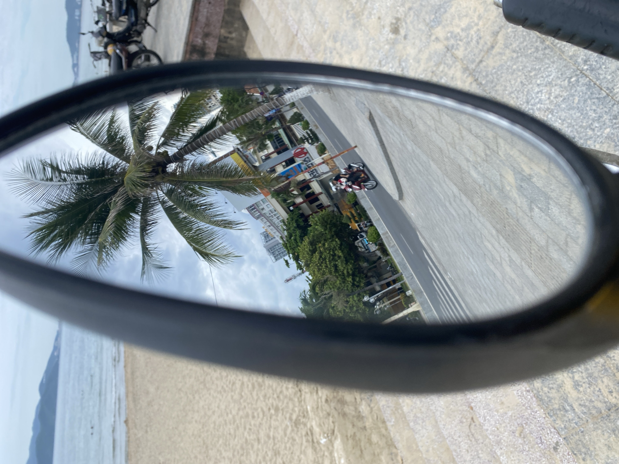 The image is a reflection in a motorcycle mirror, showing a beach and a street scene. The beach on the left is sandy with a gentle slope towards the ocean. The ocean is visible in the background with a few small figures swimming or wading. Distant hills with a light haze are visible along the horizon.
The reflection in the mirror shows a street running parallel to the beach. A person on a red scooter is riding on the street, wearing a helmet and a white shirt. To the right of the street, there is a sidewalk with patterned stone paving. Beyond the sidewalk, there are palm trees and buildings, typical of a coastal city. Some of these buildings are multi-story and appear to be hotels or apartments. A tower with antennas is also visible among the buildings. In the far right of the mirror's reflection, several parked motorcycles are visible, suggesting a public area. The sky is overcast. The overall scene evokes a tropical, urban coastal environment, consistent with Nha Trang, Vietnam.