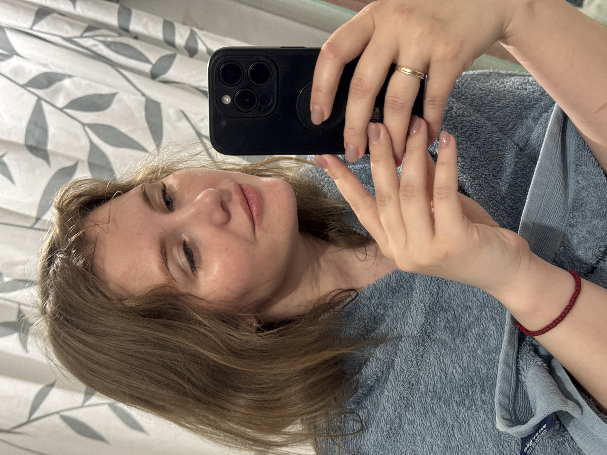 A fair-skinned person with light brown hair is holding a black smartphone in their right hand to take a selfie. The person's left hand is also visible. They are wearing a gold ring on their right ring finger and a red bracelet on their left wrist. The person appears to be wearing a blue terrycloth robe or towel. The background consists of a patterned shower curtain with a white and grey leaf design. The location is unspecified, but the prompt indicates it is potentially in Gornyi, Dvurechenskoe S/P, Russia.
