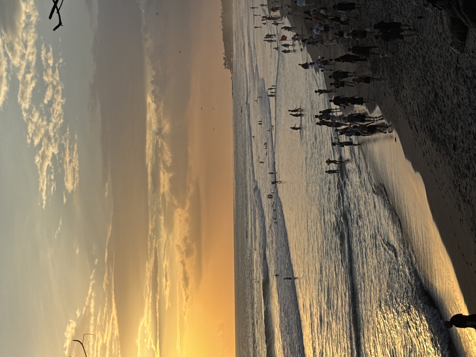 The image shows a beach at sunset in North Kuta, Indonesia. A crowd of people is near the shoreline of the beach, where gentle waves break. Some people are in the water. The sky is filled with clouds and a golden glow of sunset.