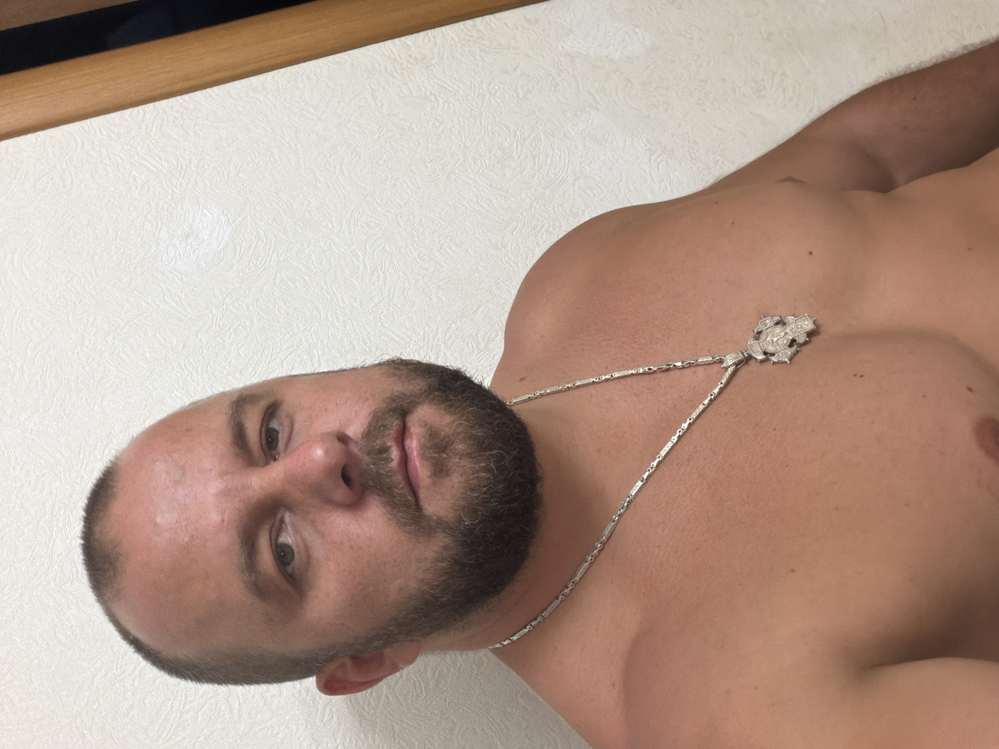 A man with a shaved head and a beard is shown from the chest up, looking towards the camera. He is wearing a silver chain with a pendant. The background is a textured white wall, with a vertical wooden element on the right side of the frame. No specific location in Sochi, Russia is discernible from the image.