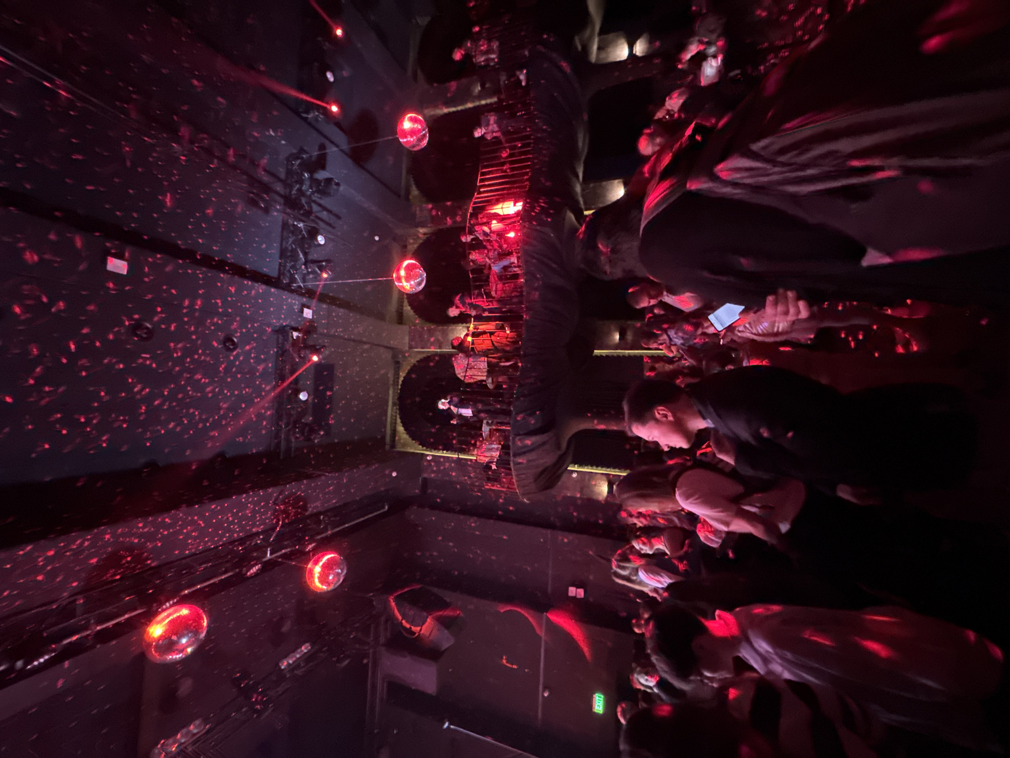The image captures a vibrant, dark-lit indoor venue, likely a concert hall, club, or bar, from an elevated perspective looking down and across multiple levels. The dominant lighting is a rich red, emanating from numerous laser projectors that create countless small, bright dots scattering across the walls, ceiling, and floors, giving a dynamic, particle-like effect. Several small, spherical disco balls are visible, reflecting and amplifying the red light.
A large crowd of people occupies various areas of the venue. In the foreground and right side, individuals are seen from above and behind, standing closely together. Many are looking towards a central area, possibly a stage or performance space, which is faintly illuminated in the mid-ground. Some individuals are seen looking down at their phones, their faces dimly lit by the screen glow.
The venue features multiple architectural levels with railings, suggesting balconies or mezzanines from which the scene is observed. On a lower wall in the mid-ground, a green "EXIT" sign with an arrow pointing right is clearly visible, indicating an emergency exit. The overall atmosphere is energetic and lively, typical of a nighttime social event or performance, with the intense red lighting setting a distinct mood. The scene conveys a sense of depth and activity within a crowded space in Sofia, Bulgaria.