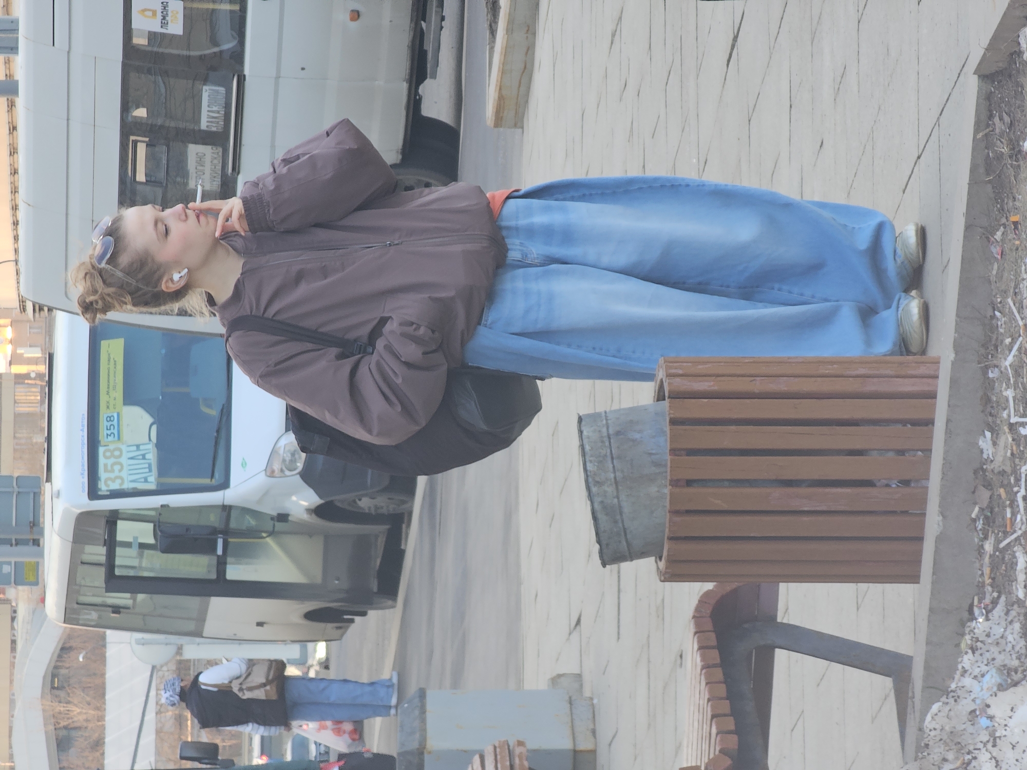 The image displays an outdoor urban scene on a paved sidewalk, identified as being in Khimki, Russia, likely near a bus stop or commercial area.
In the foreground, a young woman stands upright in profile, facing left. She has blonde hair tied in a high bun, with sunglasses resting on her head and white wireless earbuds in her ears. She wears a dark brown zippered bomber jacket, an orange undershirt visible at the waistline, baggy light-blue jeans, and light-colored flat shoes. She holds a lit cigarette between her right index and middle fingers, raised towards her mouth. A black backpack is visible on her left shoulder.
Adjacent to her, a wooden slatted trash receptacle contains a cylindrical metal bucket. To the left of the trash can, a section of a curved wooden park bench with a dark metal frame is visible. The ground is paved with light-colored square tiles. Below the paved area, a dirt strip along a curb is strewn with scattered litter, including numerous cigarette butts. A mound of dirty snow or ice mixed with debris is present on the far left.
In the mid-ground, a white GAZelle-type minibus, consistent with a marshrutka, is stopped or parked. Its front display shows the route number "358" and the destination "АШАН" (Auchan). The front passenger door is open. Cyrillic text including "ООО Красногорск-Авто" is visible above the windshield, and "ЗАКАЗНОЙ" and "ЛЕМОНА ПРО" are visible on its side windows. A second white minibus is partially visible behind the first.
Further in the background, another individual is observed from behind, wearing a white and blue patterned head covering, a dark vest over a white long-sleeved shirt, and light blue jeans. This person carries a beige shoulder bag and a white plastic shopping bag with red markings. Bare trees and distant buildings are visible under a clear sky.