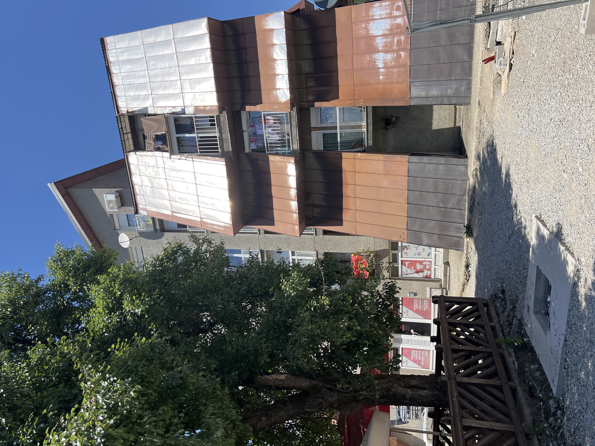 A bright, sunny day with a clear blue sky is depicted in this image, taken in the city of Mojkovac, Montenegro. The scene focuses on the exterior of an apartment building with a distinctive, modern addition clad in metallic panels of copper and silver. These panels have a weathered appearance, with streaks of rust and discoloration visible, particularly on the copper sections. The building itself has multiple stories, with visible windows and balconies. On some balconies, laundry is hung out to dry, suggesting residential occupancy.
To the left of the building, a large, leafy tree with a thick trunk provides shade. Beneath the tree, a wooden fence encloses an outdoor seating area, possibly part of a cafe or restaurant, with a red umbrella visible. In front of the fence, the ground is covered in gravel. A square concrete structure with an open center, containing some debris, is also present on the gravel.
The buildings in the background appear to be typical residential structures, with one having a satellite dish and an air conditioning unit. Further in the background, storefronts with signage are visible, suggesting a commercial area. Construction fencing and materials, including red pipes and concrete blocks, are scattered on the gravel in the foreground and to the right, indicating ongoing or recent construction work. The overall impression is of a lived-in urban environment with a blend of older and newer architectural elements.