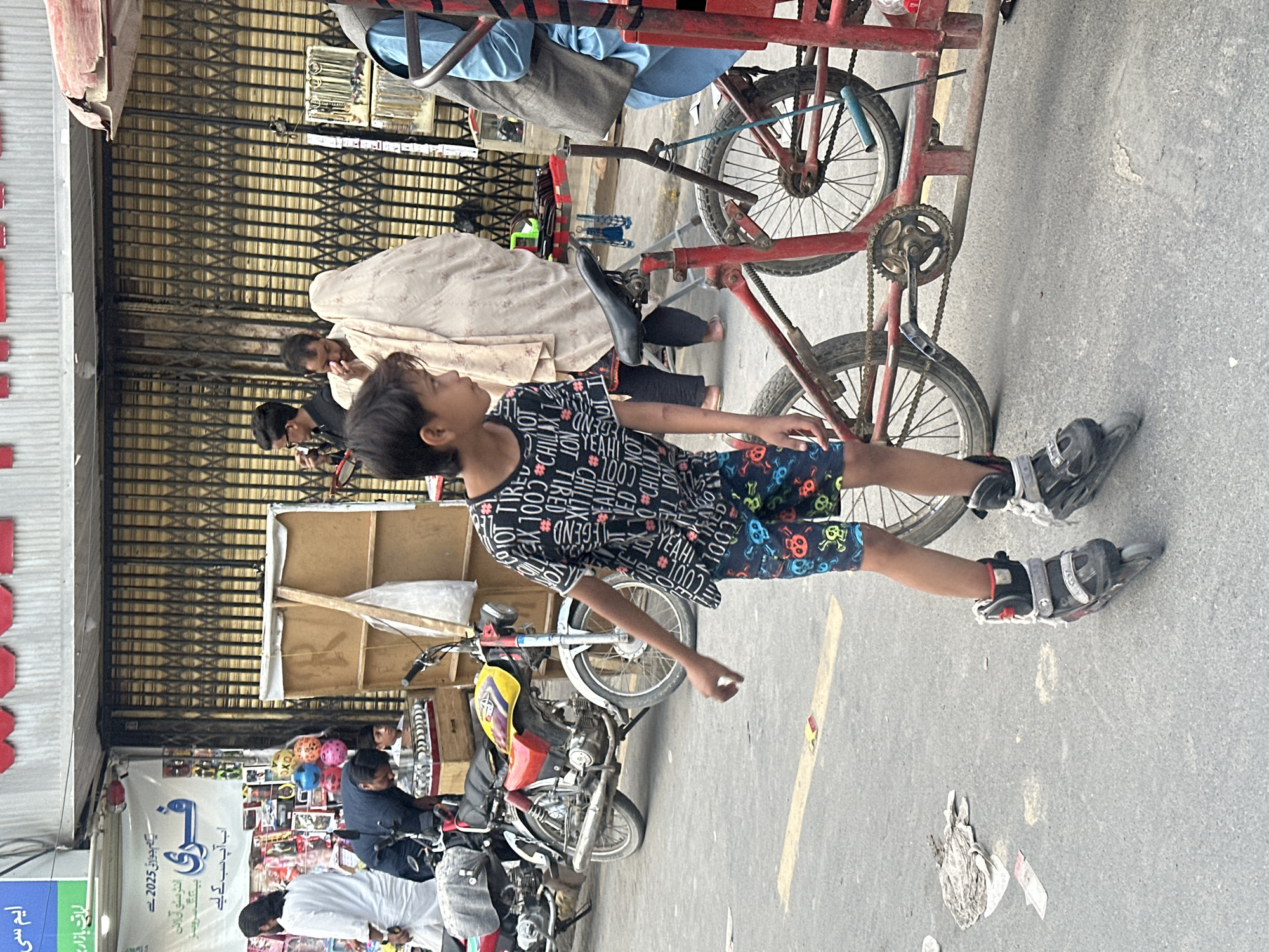 A child in roller blades stands next to a red tricycle. The child is wearing a black shirt with printed words and patterned blue shorts. Several adults are behind the child. Parked scooters and motorcycles are in the background. The location is a city street with shops. A sign in Urdu reads "Free." The sign also reads "1st July 2025."