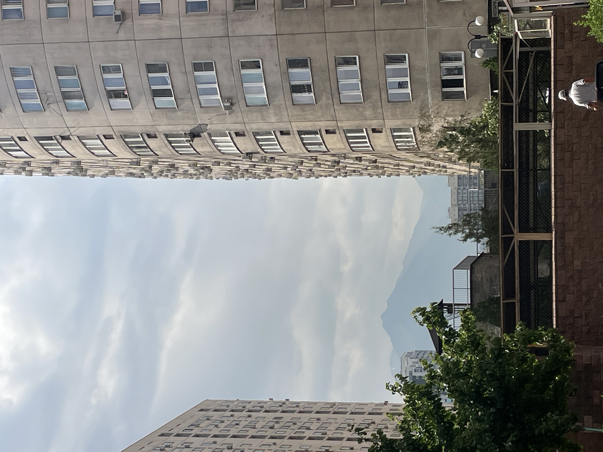 This is a daytime, eye-level, medium shot capturing a street view in Almaty, Kazakhstan, with a prominent curved residential building on the right and another apartment building partially visible on the left. The buildings feature a grid of windows and numerous external air conditioning units. In the background, mountains are visible through a hazy sky. In the foreground, a person wearing a white shirt and cap, carrying a cane, walks away from the camera towards a brick wall. Further down the street, a large tree with dark green foliage partially obscures a low-rise structure with a fenced area and a small tower. The overall scene suggests a dense urban environment with a backdrop of natural mountainous terrain.