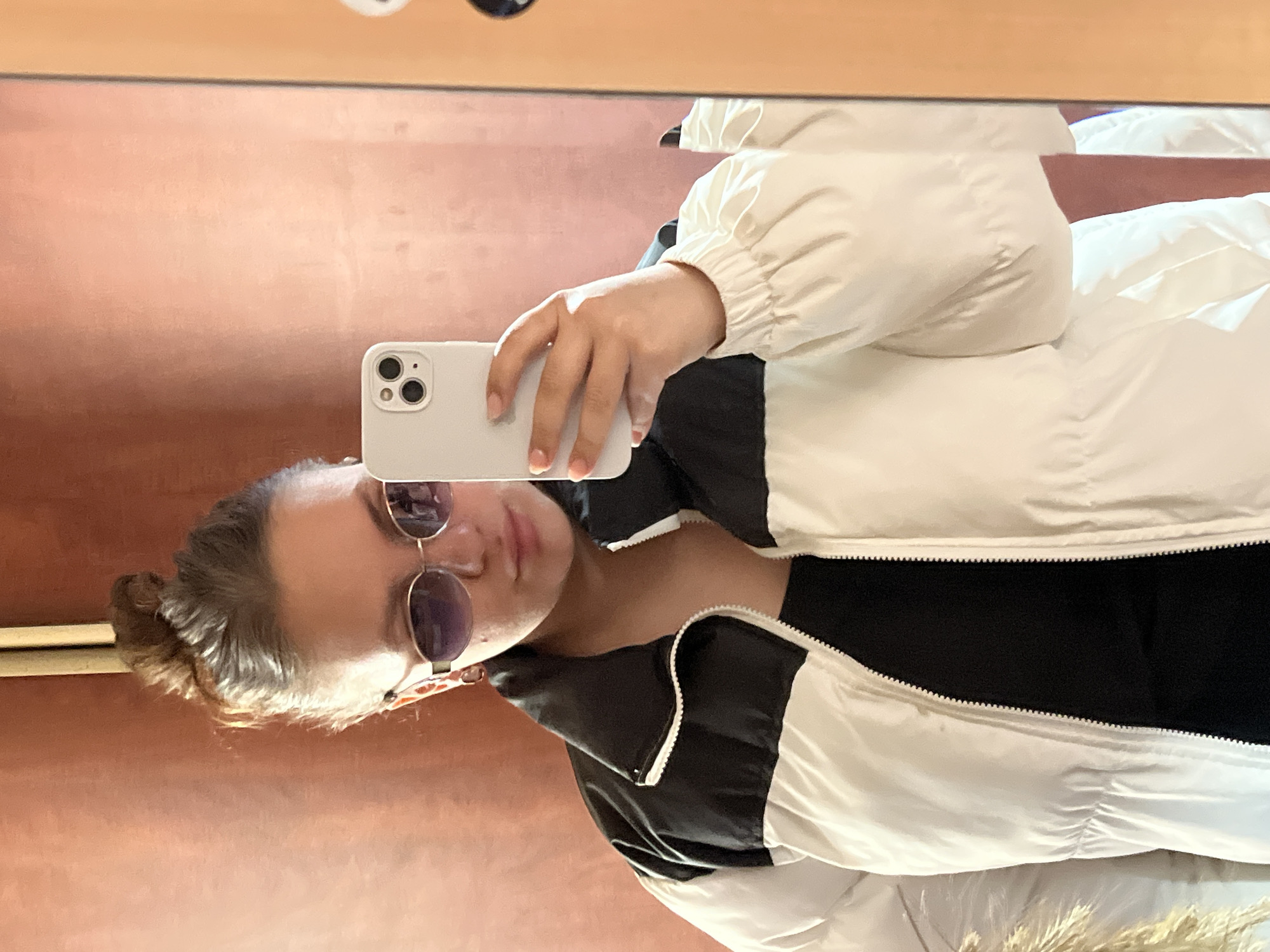 A person wearing sunglasses and a white and black jacket is taking a selfie with a white smartphone in front of a mirror. The background is a reddish-brown wall. The person has their hair in a bun. The location, Ольховка, Russia, is not discernible from the image.