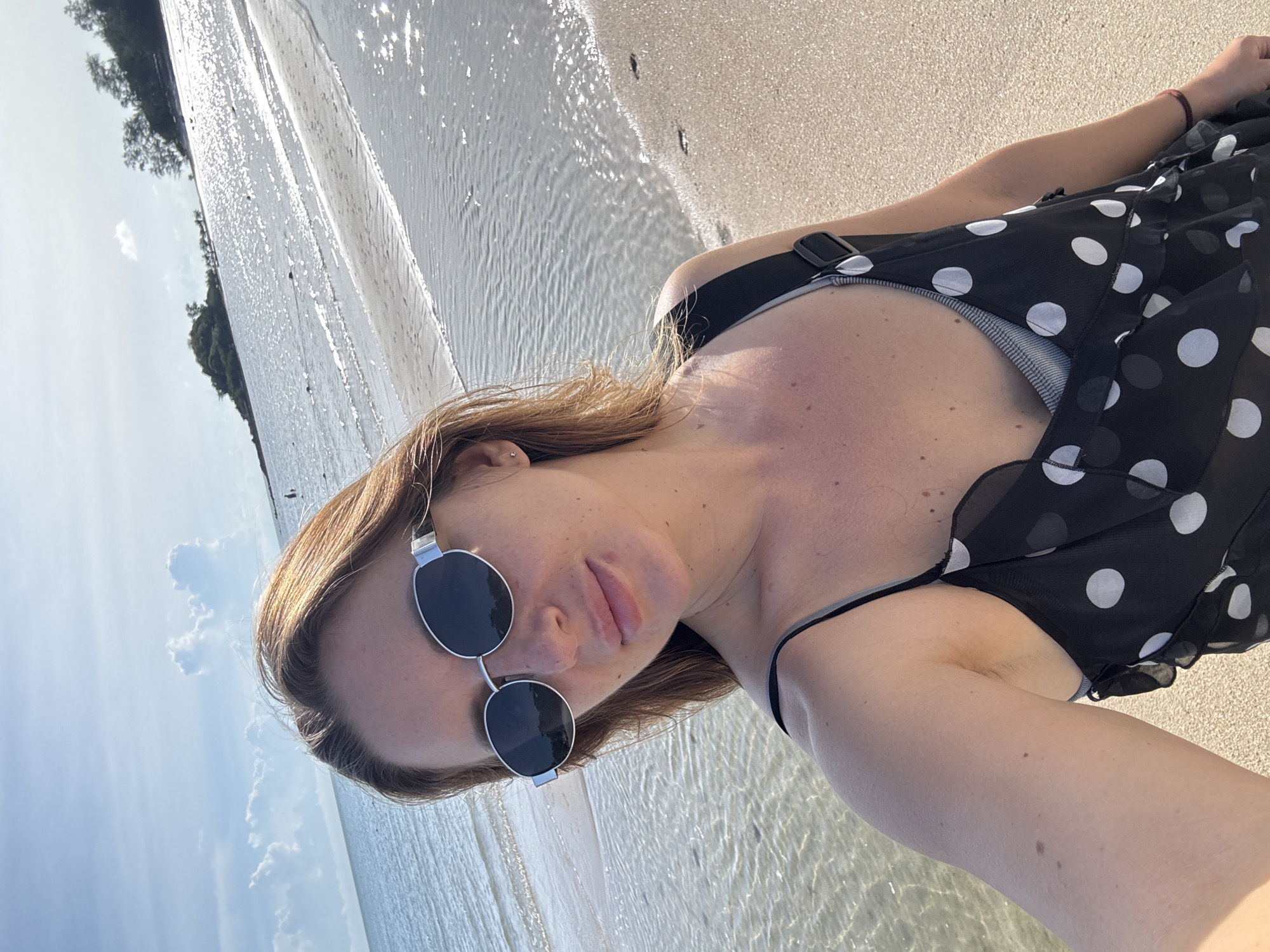 A woman is taking a selfie at the beach. She is wearing sunglasses and a black and white polka dot cover-up over a swimsuit. Her hair is long and blonde, and she has light skin. The beach is sandy, and the water is clear and shallow, with sunlight reflecting off the surface. In the background, there are trees on a hill and a blue sky with some clouds. The lighting suggests it is daytime, likely sunny. The image is oriented vertically, with the woman's head at the bottom and the beach and water extending upwards. The overall mood appears relaxed and summery. There is no visible text in the image.