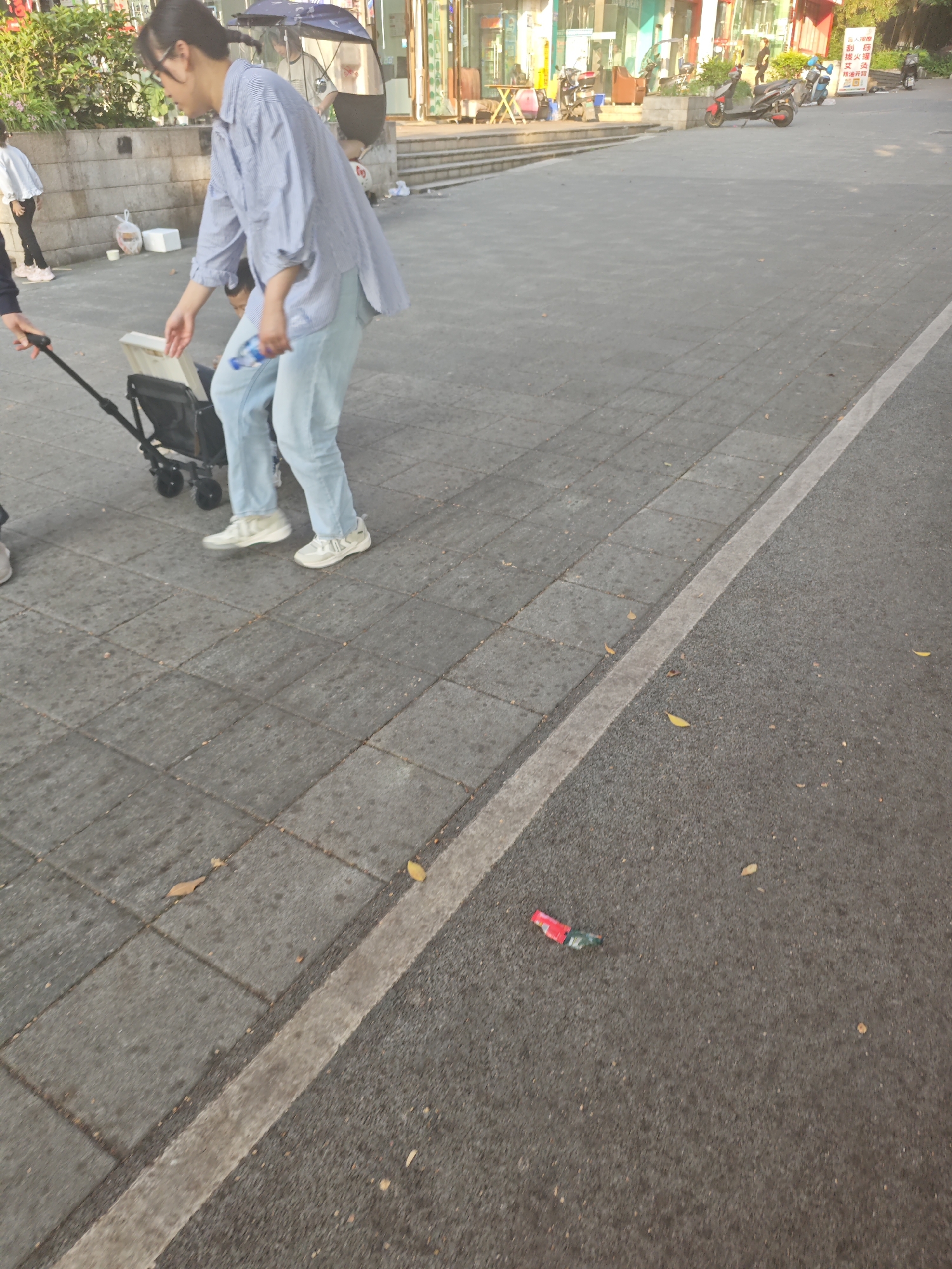 A woman with glasses and a blue striped shirt stands on a paved sidewalk next to a stroller, holding a water bottle. She is accompanied by another individual holding the stroller handle. The location is identified as Chongqing High-tech Zone, China. The pavement extends into a street section, where a piece of trash, possibly a candy wrapper, lies on the asphalt. In the background, there are buildings, motorbikes, and other people.
