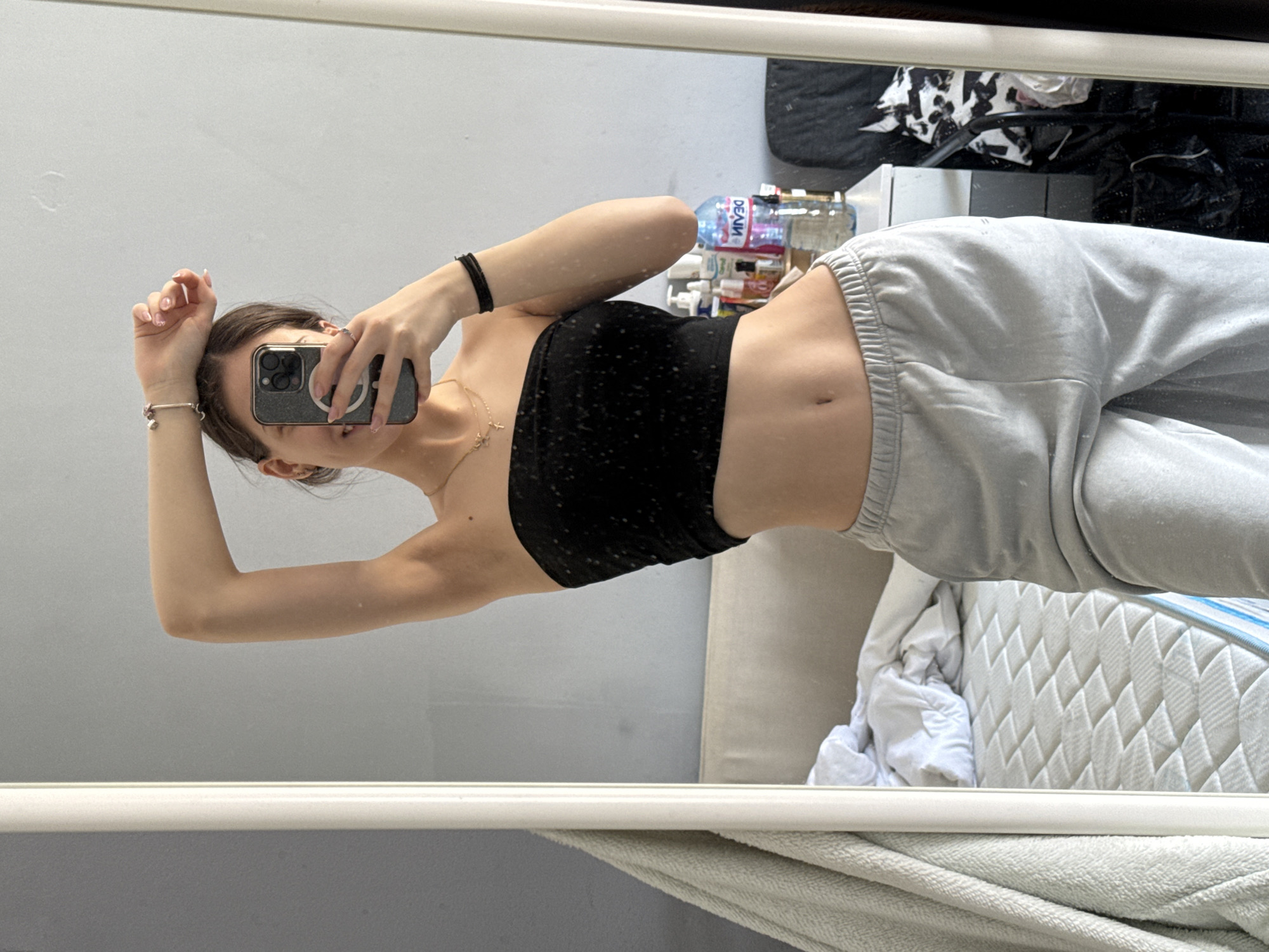 A young woman is taking a selfie in a mirror. She is wearing a black tube top and grey sweatpants. Her left arm is raised above her head. She is holding a smartphone in her right hand, obscuring her face. She has brown hair pulled back. The background includes a grey wall, a white sofa, bedding, and clutter, including various bottles. The location is not discernable from the image.