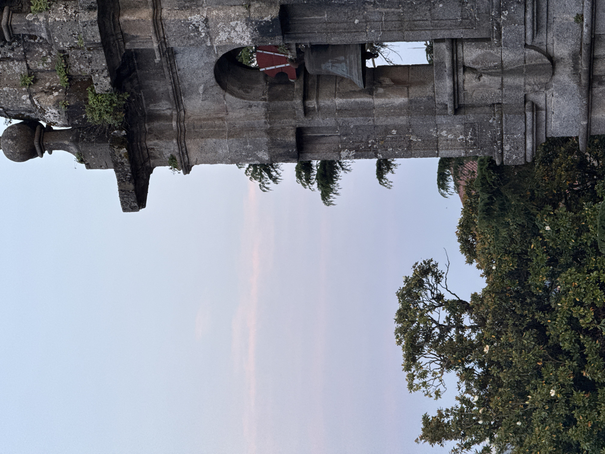 The image depicts the exterior of a weathered stone structure, likely a bell tower, against a pale sky. The structure features architectural details such as ledges, a round opening, and a bell. Some greenery grows on the stone. Adjacent to the stone structure are trees.
Although Santiago De Compostela, Spain, is mentioned, no information is provided that supports the assumption that the scene exists in that location.