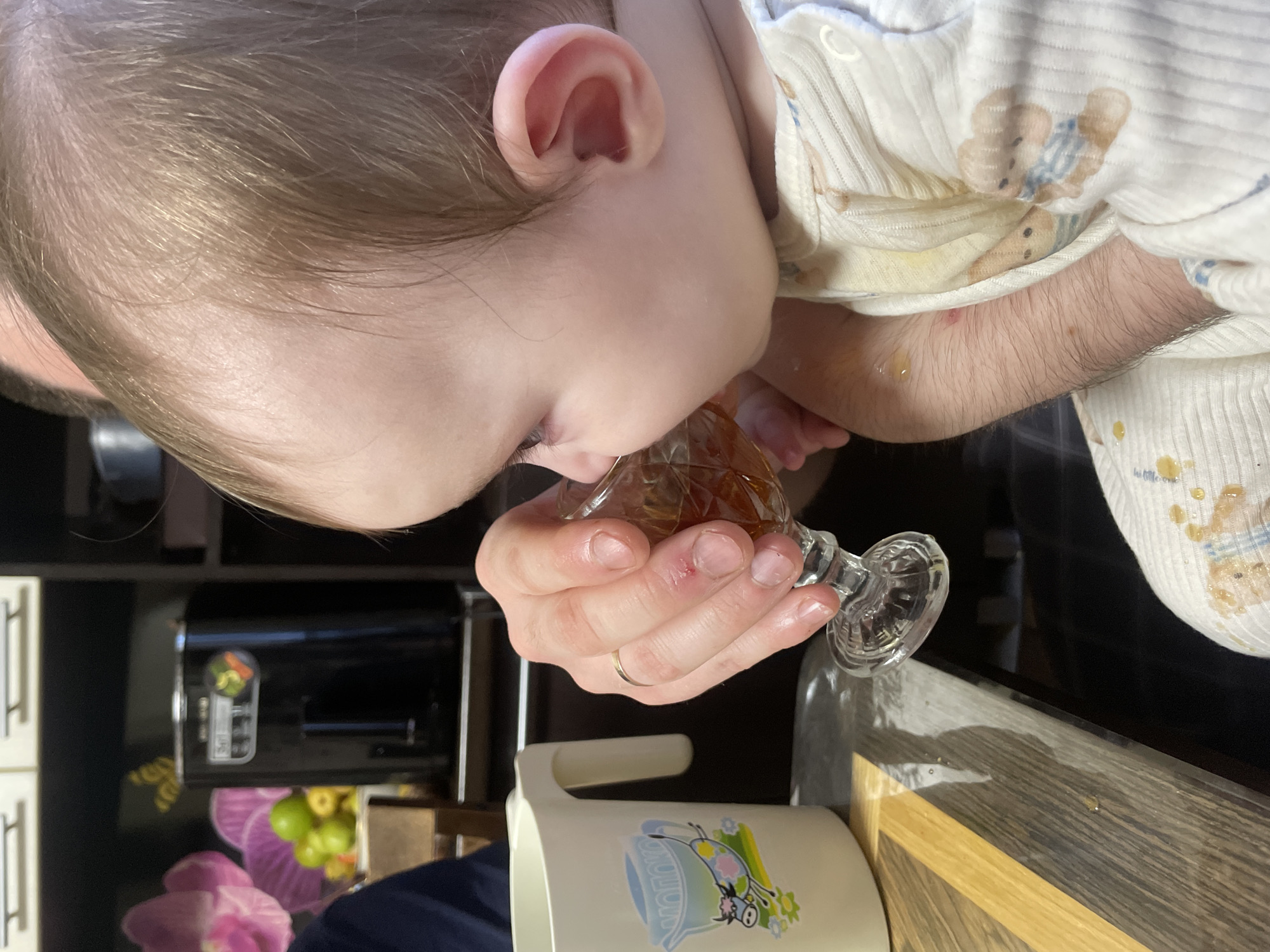A baby is drinking from a glass held by an adult. The baby is lying on its side, wearing a white patterned onesie, and has blonde hair. The adult's hand, which holds the glass, has a wedding band on the ring finger. Some liquid has spilled onto the baby’s onesie and onto the table. In the background, there is a black appliance, a decorative flower arrangement, and a white cabinet. Based on visual cues, this scene appears to be indoors, in a home setting. There is no information in the image that would indicate the location is in the city of Kuyeda, Russia.
