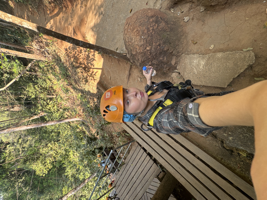 A person wearing an orange helmet and a harness stands on a wooden platform in a forest. They hold a water bottle near a large, rounded rock. The trees form a dense canopy, filtering sunlight. A zipline cable is visible stretching across the scene. The ground is a mix of dirt and stone, with exposed roots. The angle suggests a selfie taken from above.