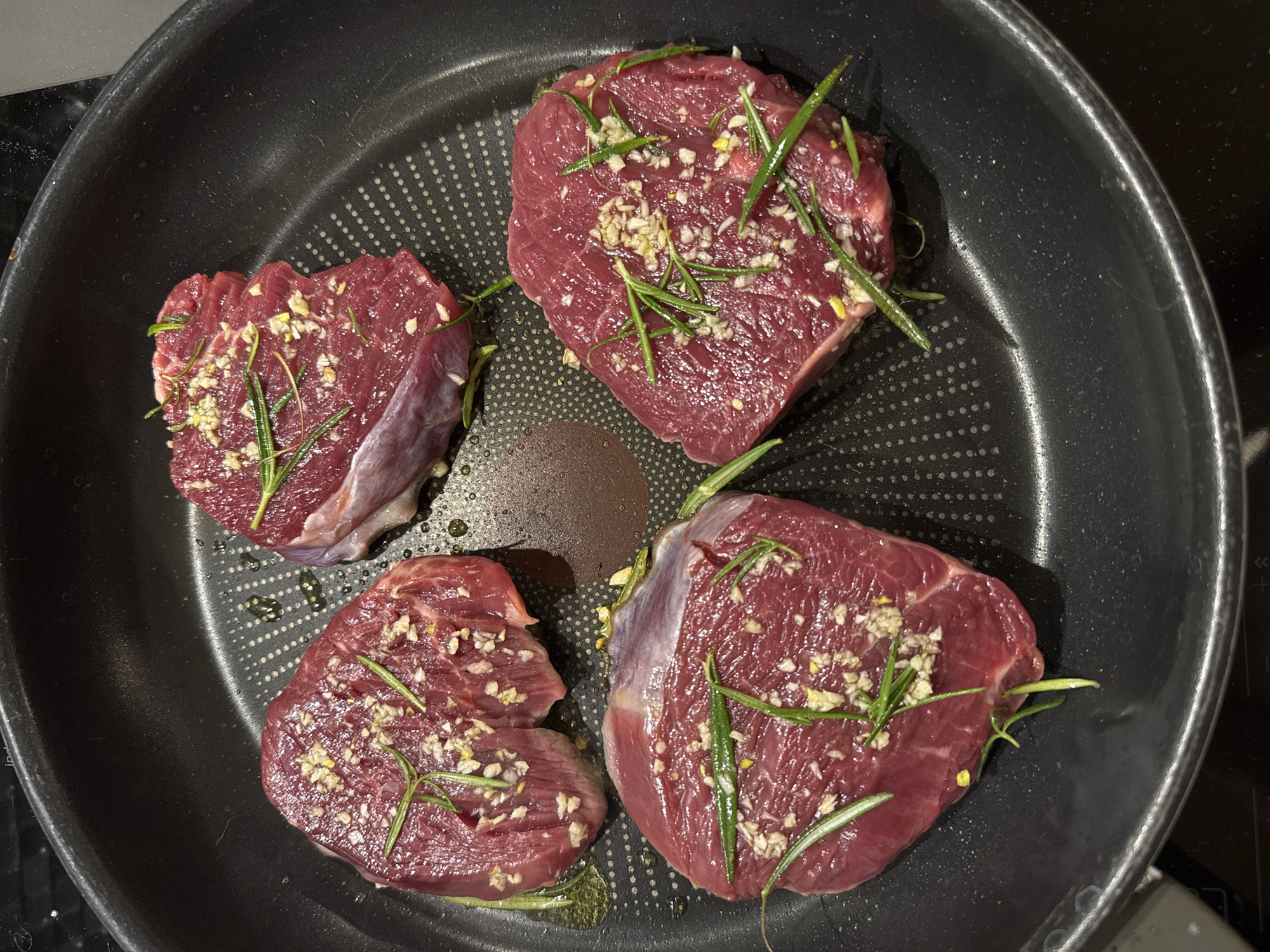 The image shows four pieces of raw, red meat, likely beef steaks, arranged in a non-stick frying pan. The meat is seasoned with chopped garlic and sprigs of rosemary. There is a small amount of oil in the pan and what appears to be burnt residue. The pan is sitting on a dark-colored stovetop. The location and city are unknown.