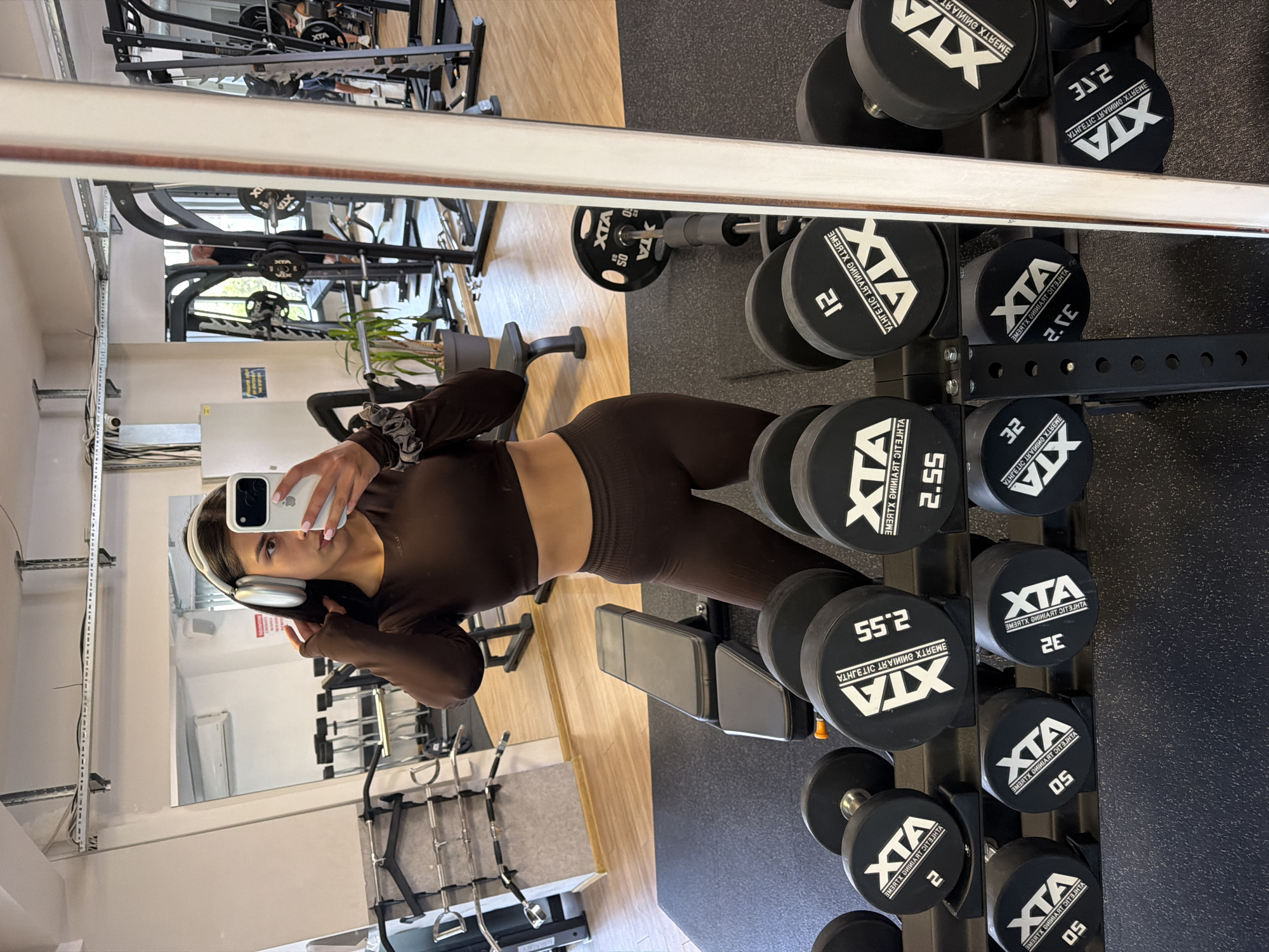 A young woman with dark hair and headphones is taking a mirror selfie in a gym. She is wearing a brown crop top and matching leggings. In the background, there are various gym equipment, including dumbbells, weight machines, and a bench. The gym appears to be indoors, with fluorescent lighting and wooden floors. The scene is clean and well-maintained. There is no indication of the location being in Gabrovo, Bulgaria.