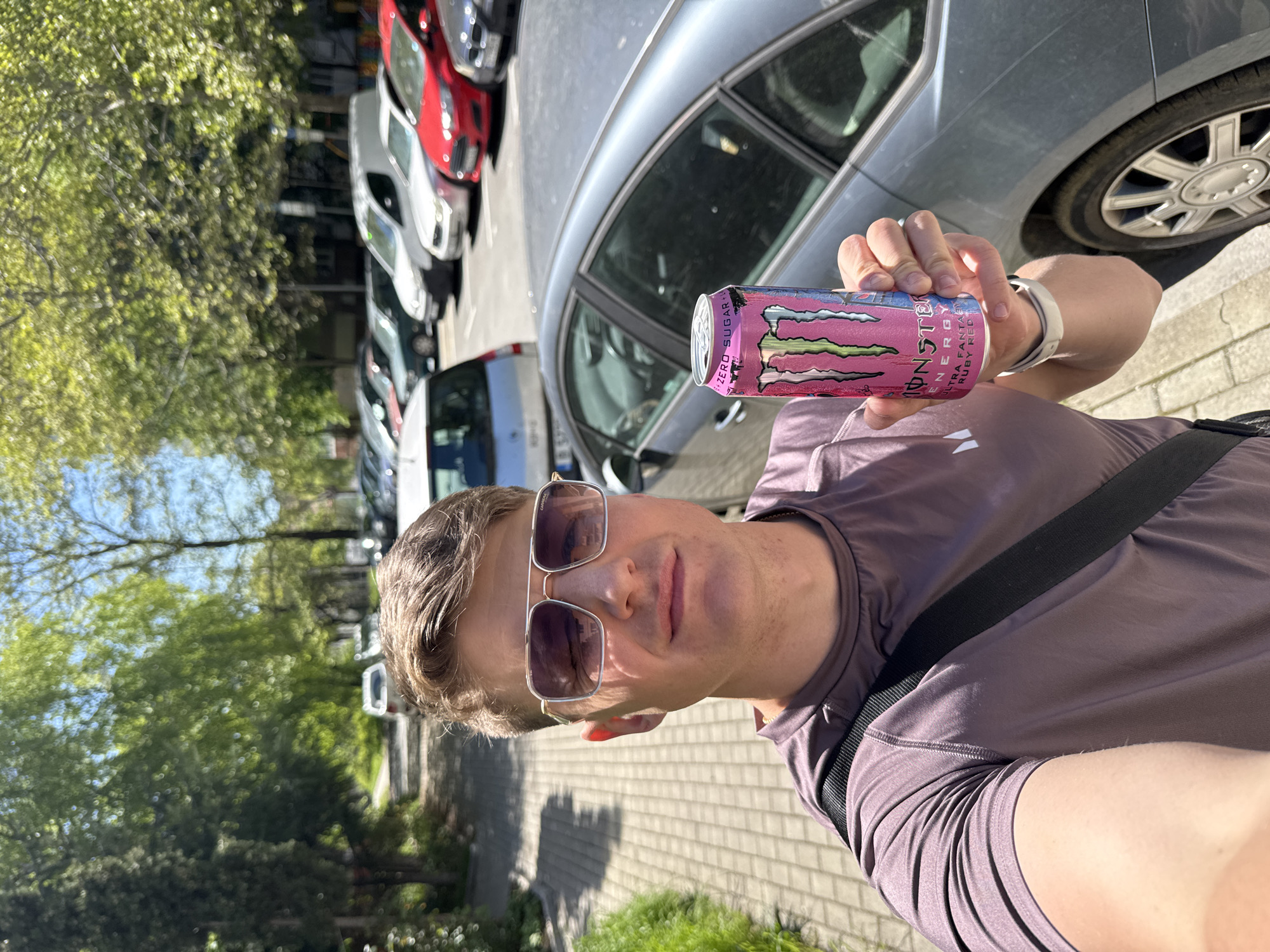 A man wearing sunglasses and a purple shirt is holding a pink Monster Energy drink in front of him. He appears to be taking a selfie. A gray car is parked next to him, and further back, there are other parked cars including a white van and a red car. There are trees and greenery in the background. The ground is paved with bricks. The specific location is indiscernible and cannot be determined as Varna, Bulgaria from the visual elements alone.