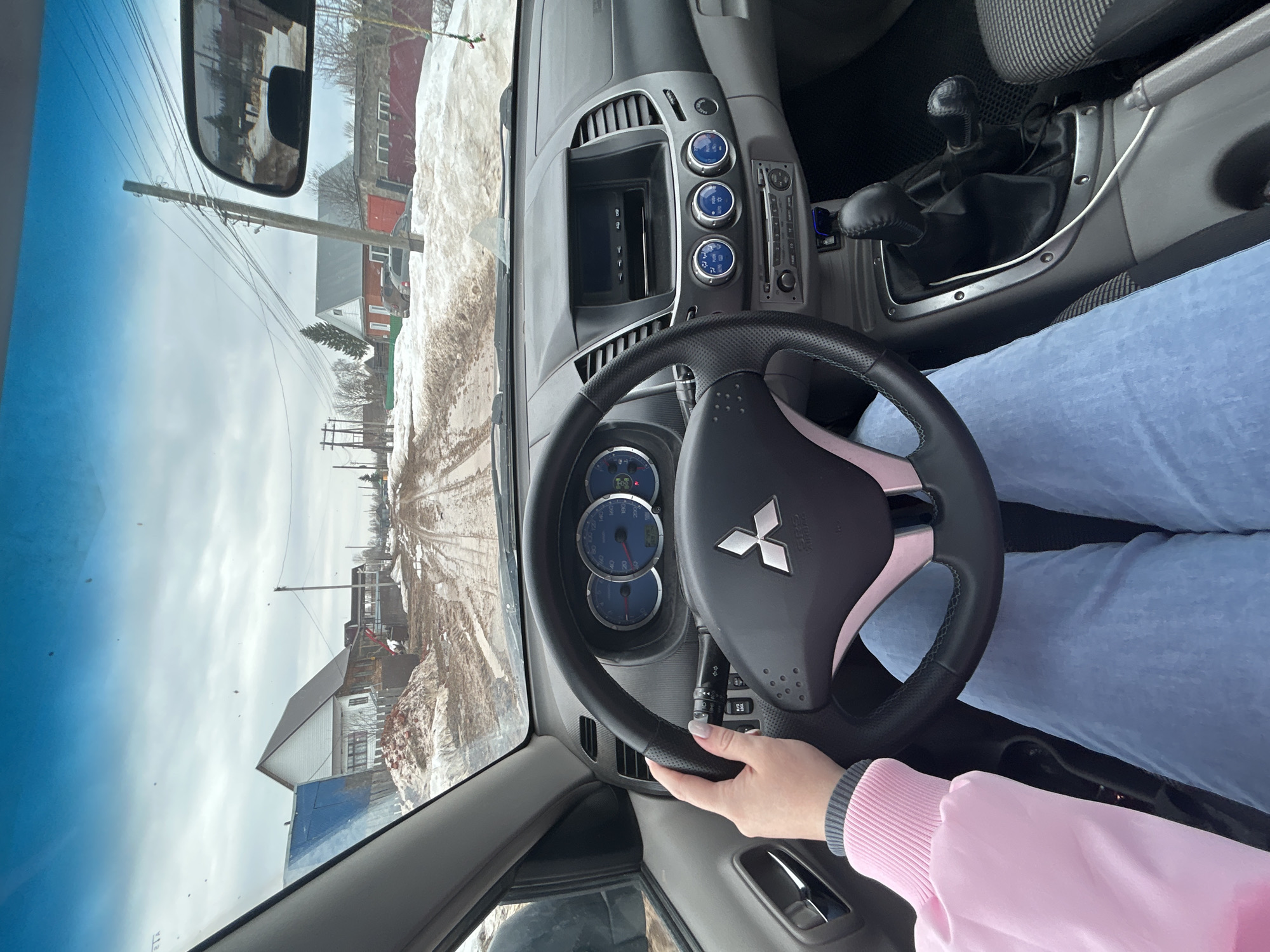 A photograph taken from within a car shows the interior, including the steering wheel bearing a Mitsubishi logo. A person wearing light blue pants and a pink long-sleeved shirt is holding the steering wheel. The dashboard features a display screen and several knobs. The gear shift is visible to the right of the steering wheel. The car appears to be on a road outside. Through the windshield, there is a view of a snow-covered street and buildings. The side mirror reflects buildings and a cloudy sky. Based on the information available online, the location is consistent with being in Фоки, Russia.