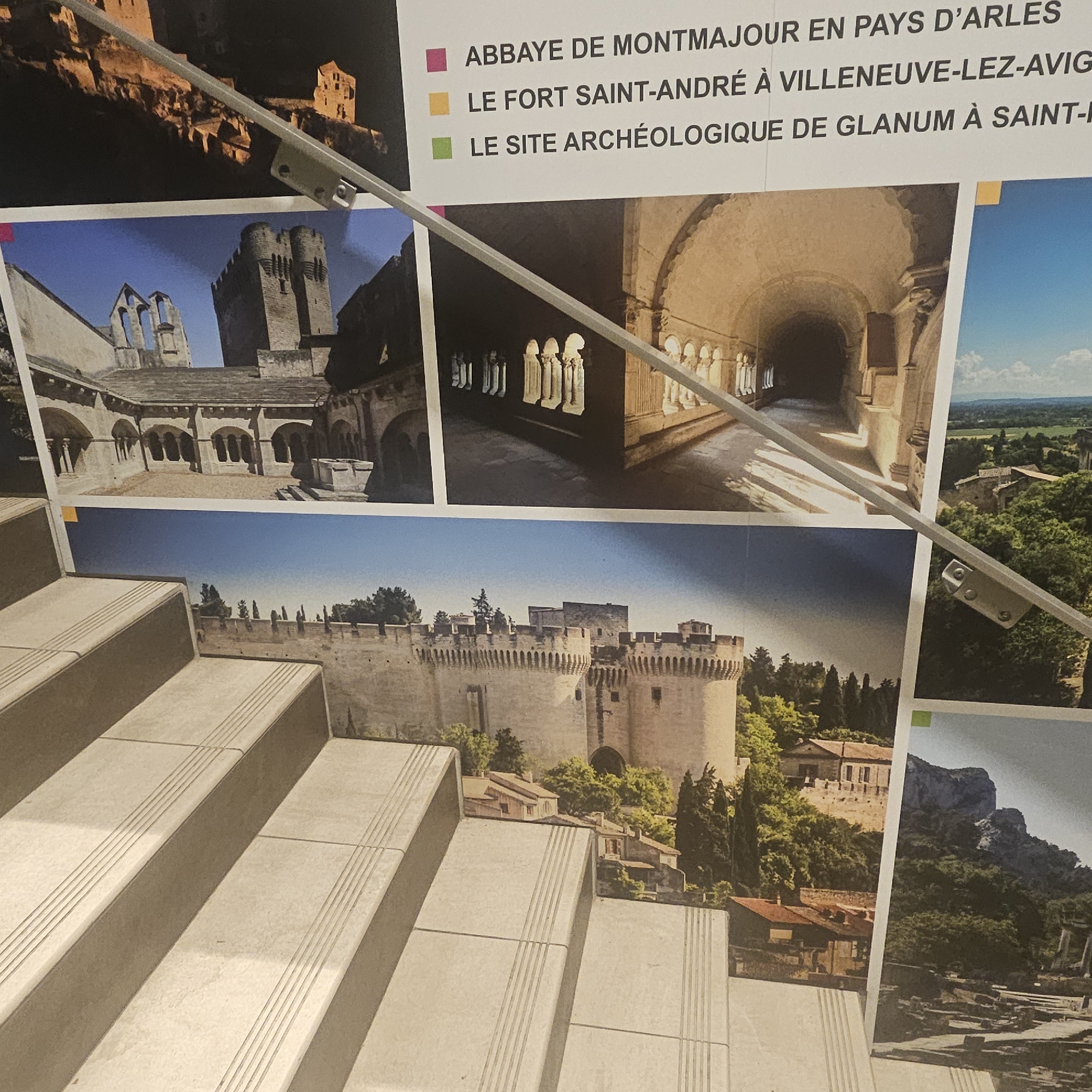 The image shows a section of a wall adorned with large photographic prints of historical sites in the region of Arles, France. A silver metal railing runs diagonally across the image, obscuring parts of the photographs and the stairwell on which the railing is mounted. The photographs depict architectural ruins and fortified structures. One print features the Abbey of Montmajour, an abbey on the outskirts of Arles. Another shows Fort Saint-André in Villeneuve-lez-Avignon. A third displays the archaeological site of Glanum in Saint-Rémy-de-Provence. The stairwell is constructed of light-colored, possibly stone or concrete, steps with a textured pattern on the risers. The overall impression is of a display in a public space, possibly a museum or tourist information center, showcasing local heritage. There are no people visible in the image.