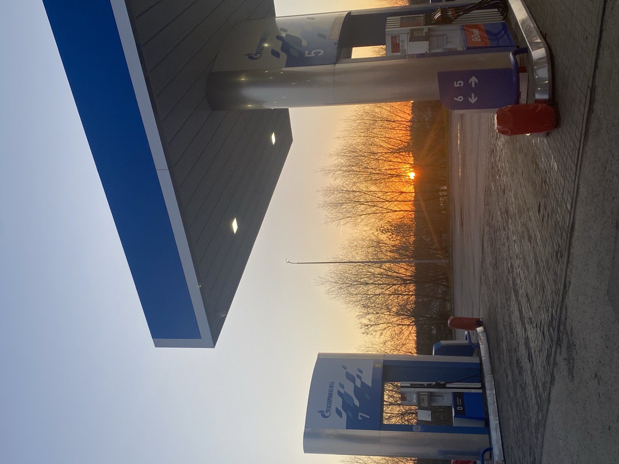 The photograph shows a Gazprom Neft gas station in Kazach'ya Sloboda, Russia, at sunset. The sun is setting behind a line of bare trees in the distance. The foregound of the shot shows a section of the station's canopy and two gas pumps. The pumps are numbered "5" and "7" in blue. Next to one of the pumps, there's a trash can and a sign indicating the pump number. The ground is paved with interlocking blocks. The sky is transitioning from a light blue to a warm orange color near the horizon.
