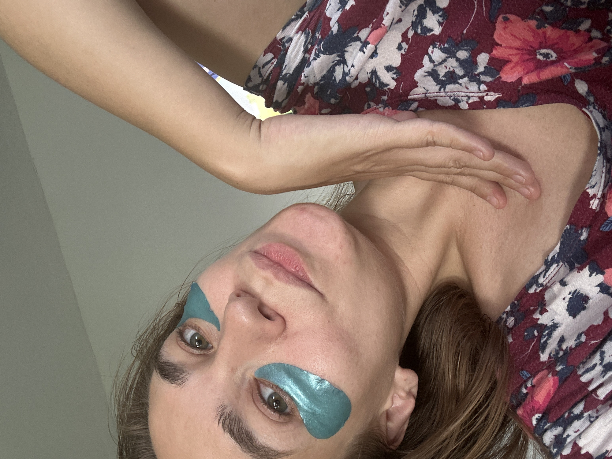 A close-up, waist-up shot shows a person with brown hair and brown eyes wearing teal under-eye patches. Their left arm is bent and resting on their chest, and their right arm is partially visible, positioned against their neck. They are wearing a maroon floral-patterned top. The background is a plain, light-colored wall. The scene appears to be indoors.