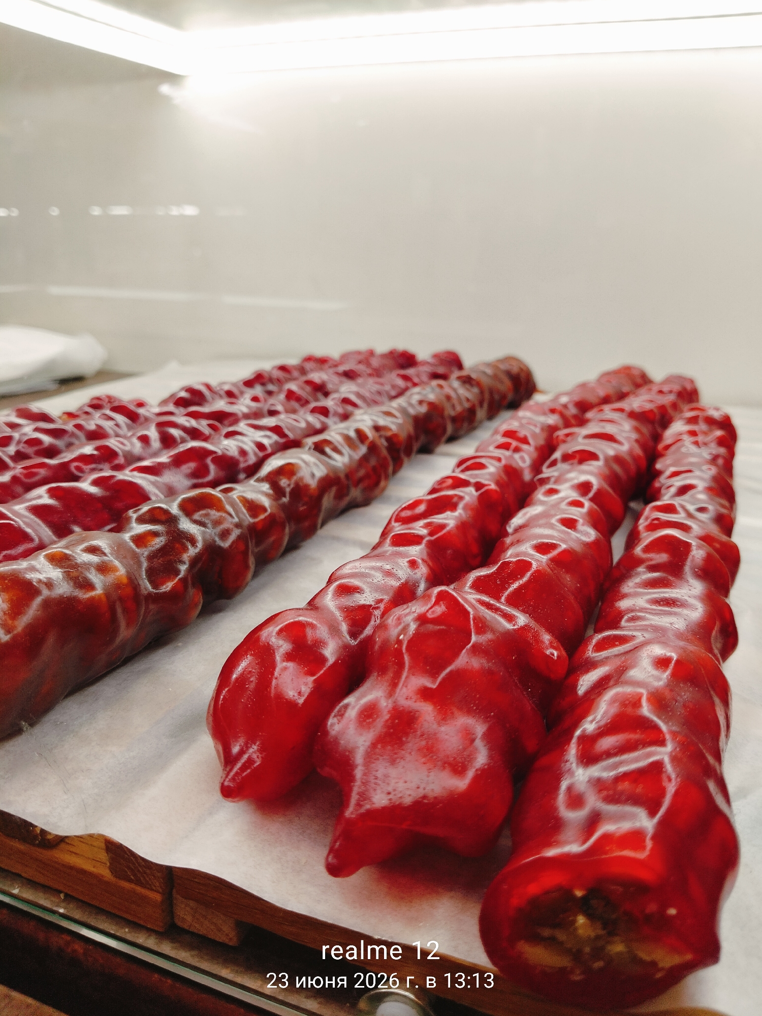 This image shows a close-up, eye-level view of multiple strings of churchkhela, a traditional Georgian candy made from grape juice and nuts, arranged on a white surface. The churchkhela are uniformly red and appear to be coated in a glossy, transparent shell, with some strings showing exposed nuts within. The lighting is bright and overhead, creating reflections on the candy's surface. In the bottom right corner, text indicates "realme 12" and the date "23 июня 2026 г. в 13:13", suggesting the photo was taken with a Realme 12 phone at 13:13 on June 23, 2026. The context of Shchyolkovo, Russia, is not visually represented in the image.